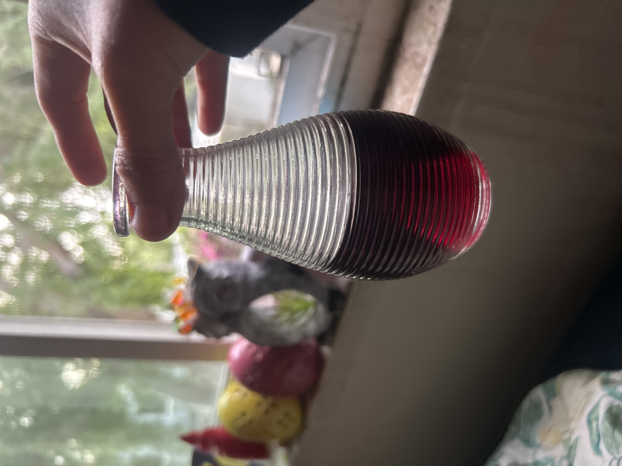 A person's hand is holding a clear, ribbed glass vase filled with a red liquid. The vase is held in front of a window with an outdoor view. On the window sill, there's a stacked ornament of a cat, a grey-green sphere, a pink sphere, and a yellow sphere. The image is taken indoors, with a wall to the right and part of a bed visible in the lower right corner. The location is implied to be in Los Angeles, United States of America based on the prompt's instructions.