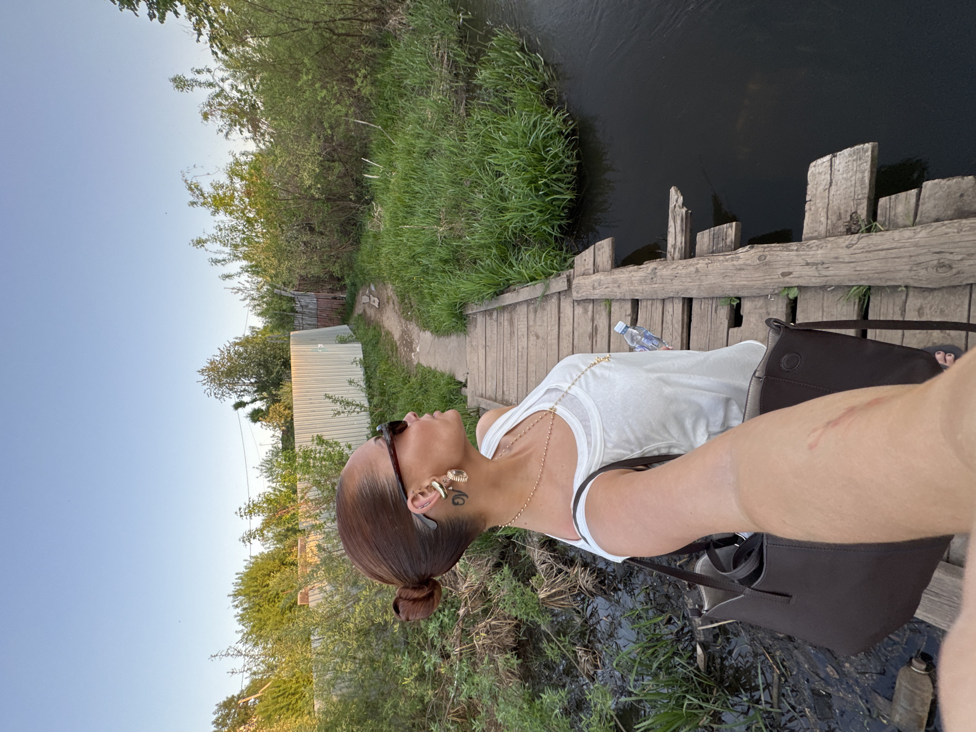 A woman is taking a selfie on a wooden bridge in a rural-looking area. The bridge crosses a narrow body of water, possibly a stream or small river. The surrounding environment includes green vegetation and some trees. A fence can be seen in the background. A water bottle is visible on the bridge. The woman has a dark purse and is wearing a white shirt, jewelry, and sunglasses. Based on the rural setting and style, it is not possible to determine the exact location in Chaikovsky, Russia without more context or specific landmarks.