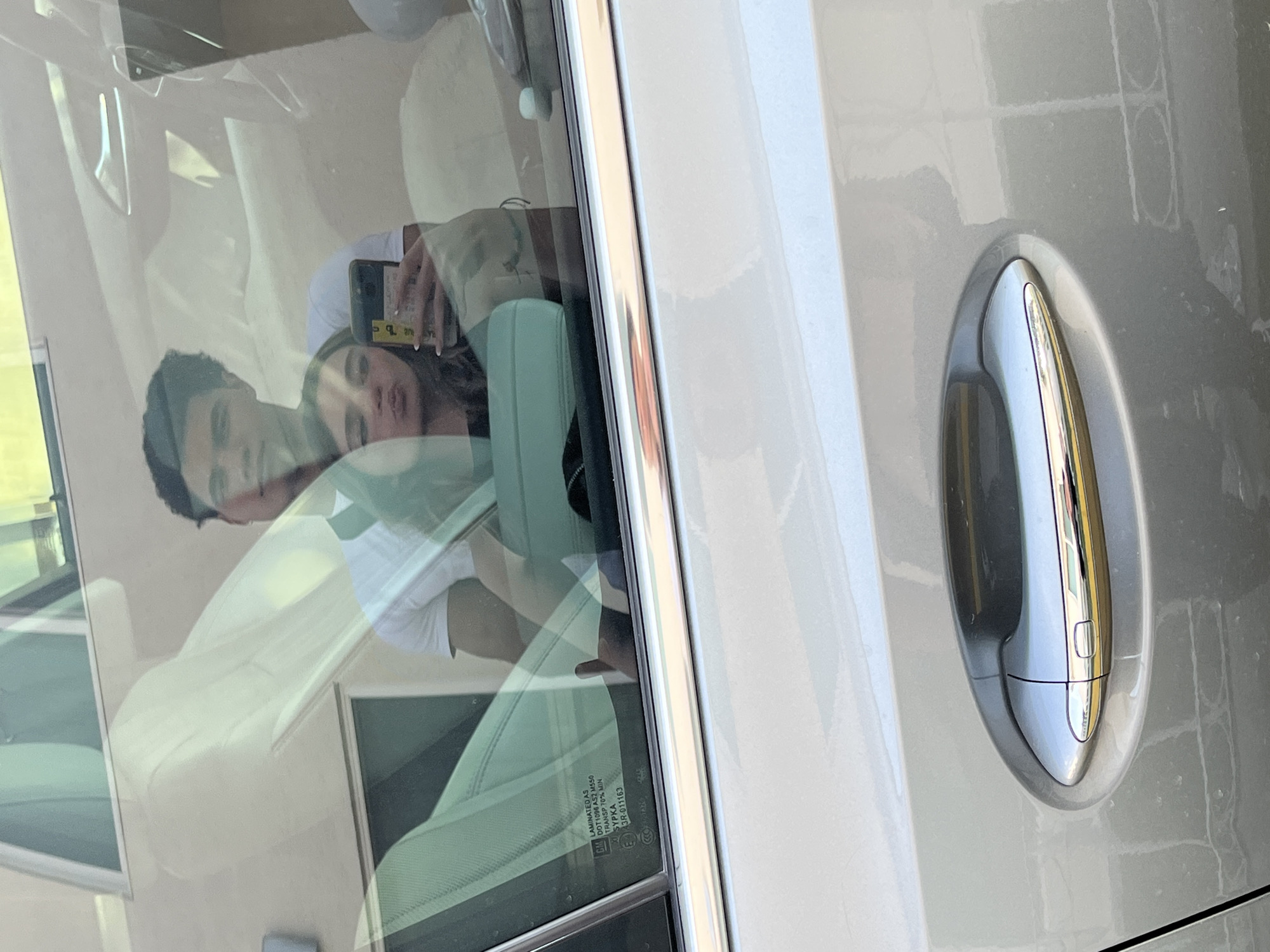 A couple is reflected in the window of a parked car. The man is on the left, wearing a white t-shirt. The woman is on the right, holding a mobile phone, and the camera is directed towards them. The vehicle door handle is in the foreground. The reflection is distorted by the glass of the car window. Visible on the window is the text "LAMINATED AS DOT 1096 AS2 M550 TRANSP. 70% MIN SYPKA 3R-011163". The scene is presumably set in San Bernardino Tlaxcalancingo, Mexico, although there is no direct evidence of this in the visual data.