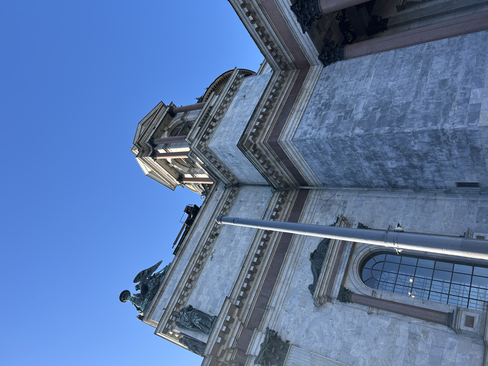The image shows a low-angle view of a building, likely a cathedral or grand structure, against a clear blue sky. The architectural style features marble or light-colored stone blocks with darker red-brown accents. Details include ornate window frames, statues of angels or figures along the facade, and Corinthian columns. A metal pole or structure is visible in the foreground, possibly part of maintenance or repair work. The building's design appears to be neoclassical or baroque revival. Given the architectural style, the location might be St. Petersburg, Russia, potentially featuring Saint Isaac's Cathedral. The scene is devoid of people.