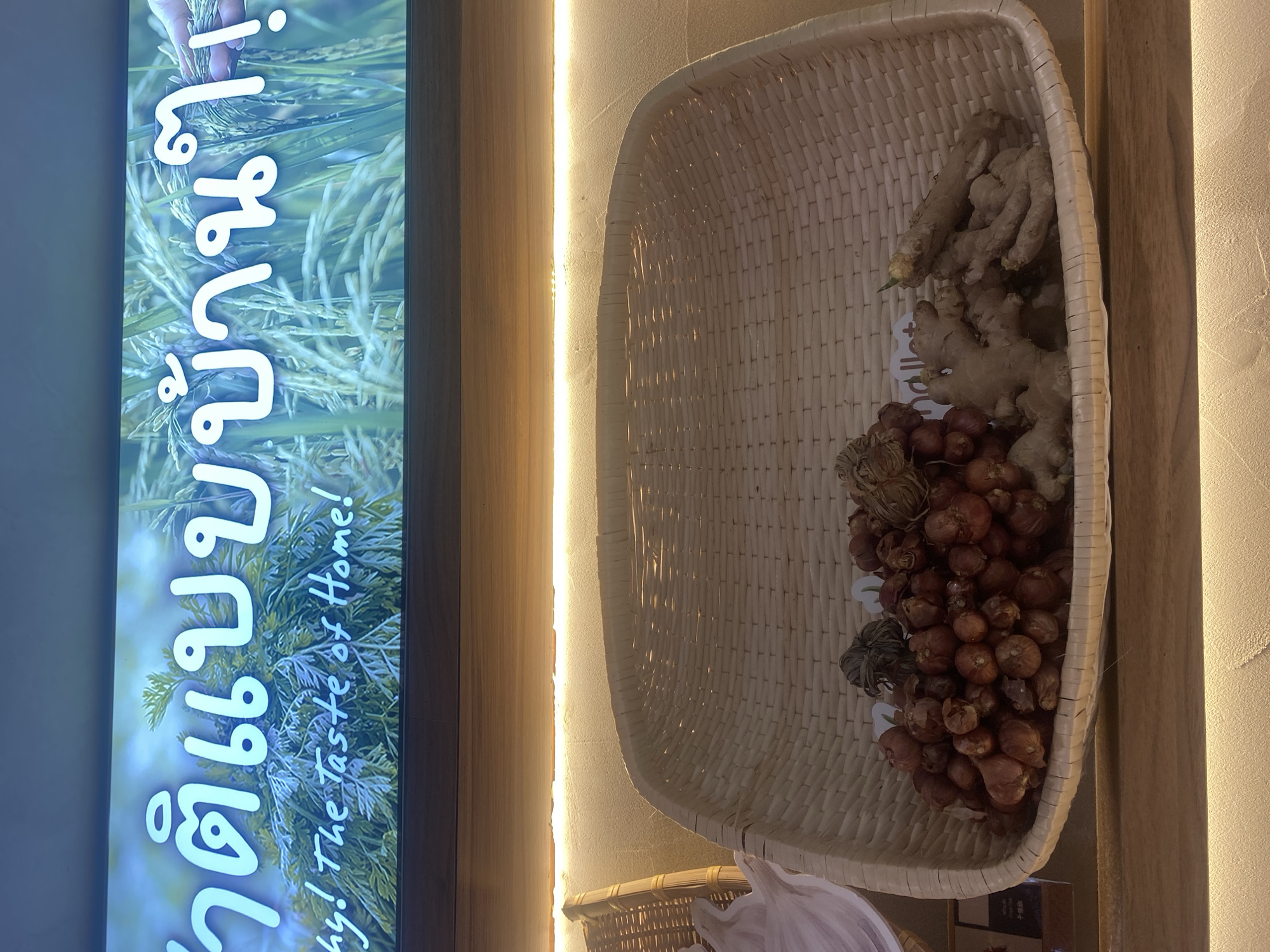 The image shows a display in Bangkok, Thailand. On the left, there is a sign with writing in Thai and English that says "The Taste of Home!". To the right of the sign, there is a woven basket filled with ginger and shallots. The display is lit with warm lighting.