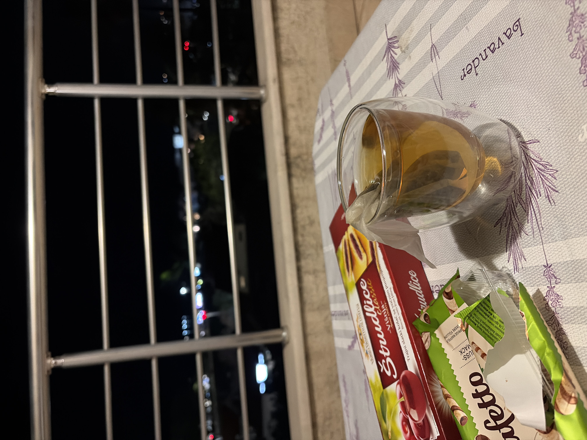 This is an indoor, medium shot, eye-level photograph taken at night from a balcony in Budva, Montenegro. The scene depicts a small table set with snacks and a drink. A glass of tea with a tea bag steeping is in the foreground, to the right. Next to it, to the left, is a red box of "Strudlice classic" biscuits with sour cherry filling, and partially unwrapped green packaging of "Afetto" wafer rolls. The table is covered with a striped cloth with a lavender motif. In the background, through a balcony railing, city lights and blurred bokeh are visible against the dark night sky.