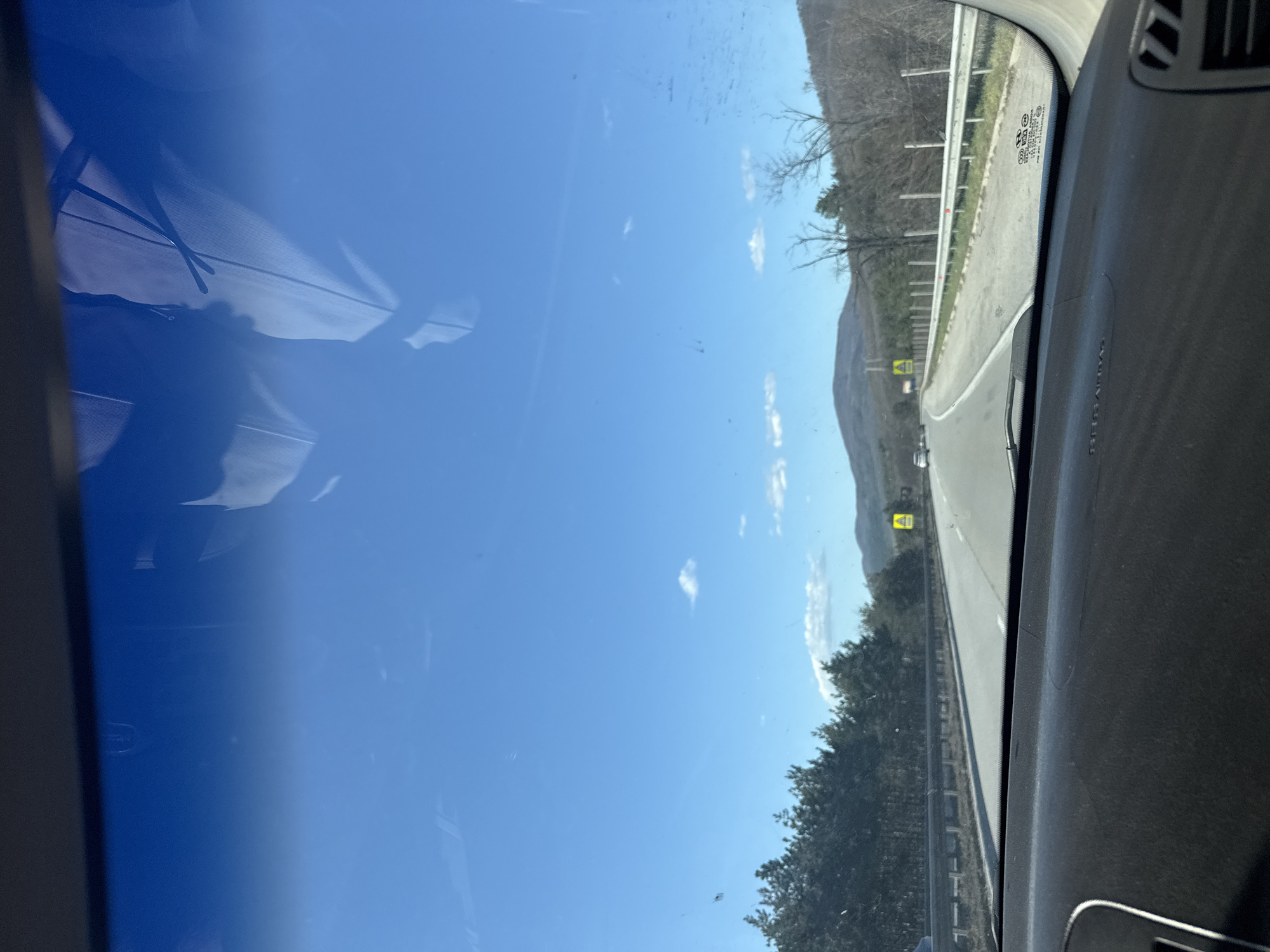 A view from inside a vehicle driving on a highway near Gorna Vasilitsa, Bulgaria. The highway is adjacent to a tree-lined area and a mountain range in the background under a clear blue sky with scattered clouds. The dashboard of the vehicle is visible in the foreground.