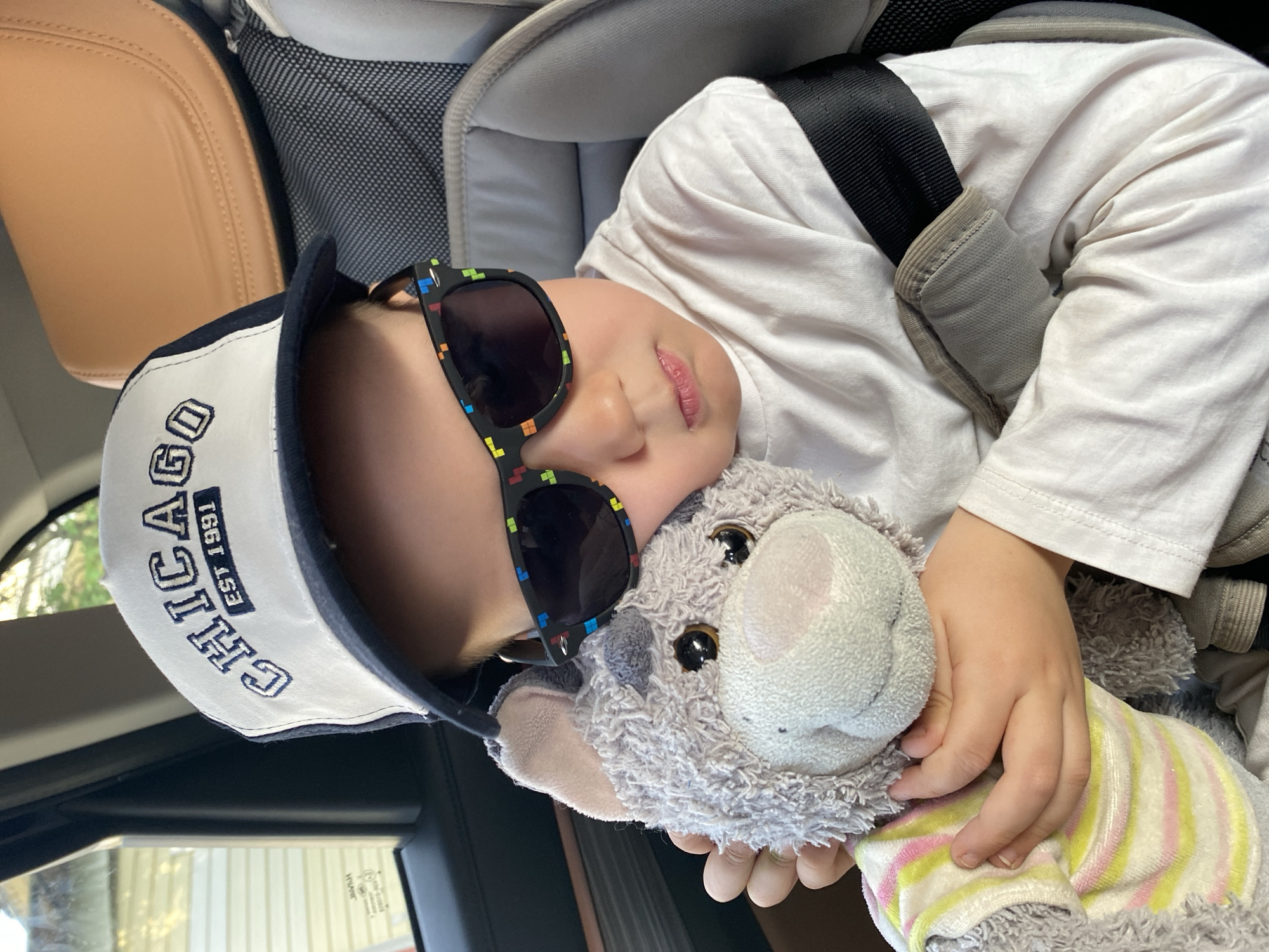 A young child, wearing a white long-sleeved shirt, a baseball cap with "CHICAGO EST 1991" embroidered on it, and sunglasses with colorful pixelated patterns on the frames, is asleep in a car seat. The child is holding a plush, gray animal toy wearing a striped shirt. A black seat belt is fastened across the child's chest. The car interior includes a tan leather seat and a gray headrest for the car seat. The child is presumably in a car, but the location of Noviy, Russia is not visually discernible.