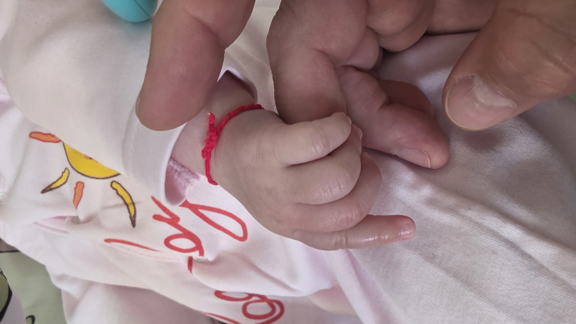 A close-up photograph depicts a baby's hand clutching an adult's finger. The baby is wearing a pink and white onesie with a cartoon sun design. A red string bracelet is tied around the baby's wrist. The adult's hand is visible, with clean fingernails. The background is a slightly blurred white fabric. There is no information available in the image to confirm the location in Blagoevgrad, Bulgaria.
