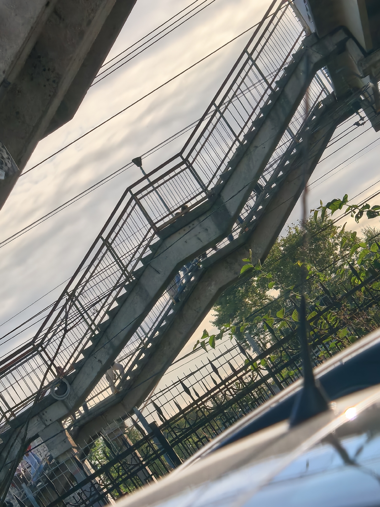 The image depicts an elevated concrete and metal pedestrian overpass, likely part of the infrastructure in Ulyanovka, Russia, given the context provided. The overpass is angled diagonally across the frame, with a clear sky and clouds visible above. Power lines are strung across the scene, intersecting with the overpass structure. To the right of the overpass, dense green foliage and trees are present, partially obscuring a black metal fence. In the foreground, the blurred surface of a car, including its antenna, is visible, suggesting the vantage point of the photographer. There are a few indistinct human figures visible on the overpass itself, though their actions and details are not clear. The lighting suggests it is either dawn or dusk, with a soft, diffused light illuminating the scene.