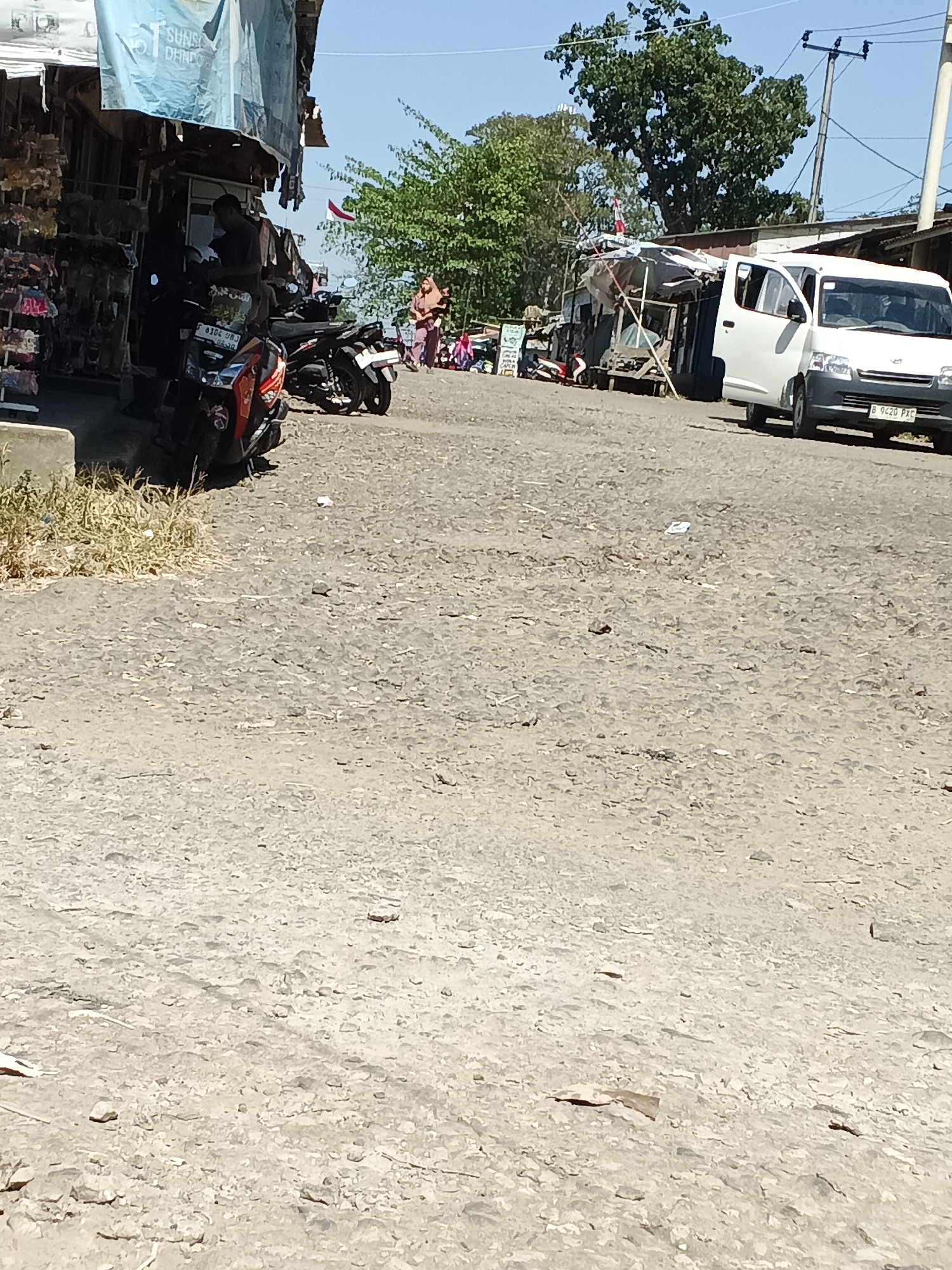 The image depicts a street scene in Warungkiara, Indonesia, on a clear, sunny day. The foreground is dominated by a rough, gravelly road surface. In the middle ground, to the left, is a market stall with various items displayed, including what appear to be sandals or flip-flops. A man is standing in front of the stall, seemingly interacting with a motorcycle. Two motorcycles are parked near the stall. Further down the road, to the right, a white van with its door open is parked. Behind the market stalls and vehicles, there are trees and a clear blue sky. A few people are visible in the background, walking or standing near the market area. A red and white Indonesian flag is flying from a pole. Power lines and utility poles are also visible in the background, suggesting a semi-urban or rural setting. The overall impression is that of a busy market or roadside commercial area.
