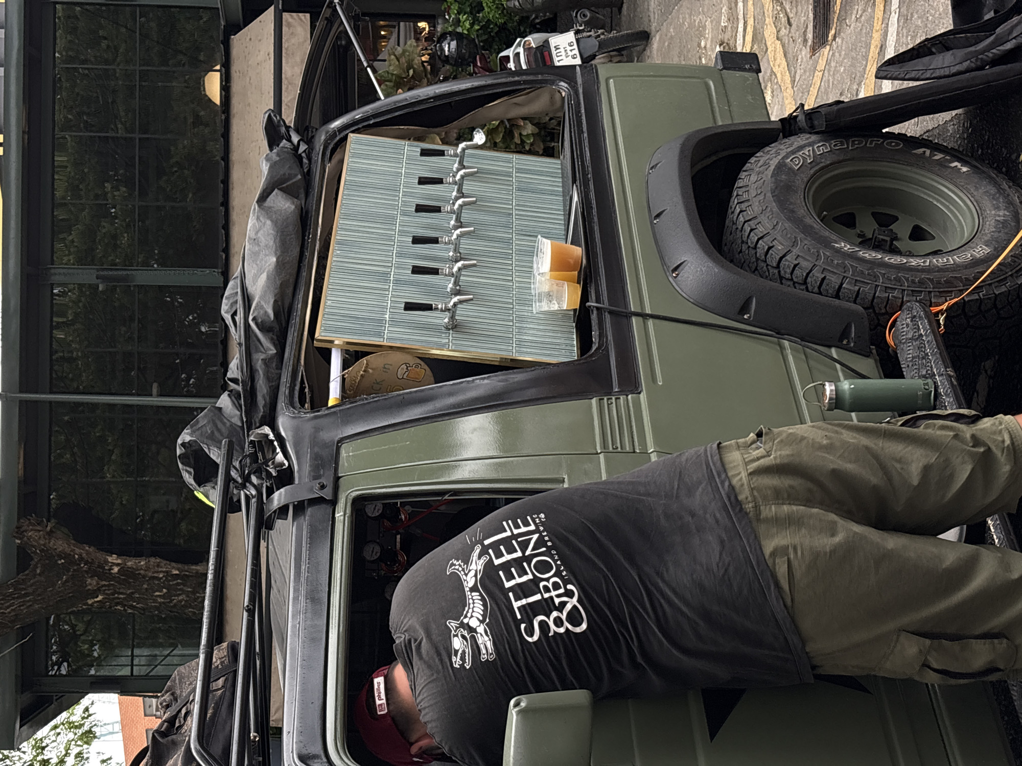 In Choeng Thale, Thailand, a person wearing a black t-shirt with a white graphic of a skeletal dog and the words "STEEL & BONE ISLAND BREWING" and olive green cargo shorts is leaning into the open rear section of a muted green, four-door SUV. The SUV has a roof rack with black bags secured to it. The rear of the SUV has been converted into a makeshift bar with six beer taps mounted on a light blue tiled panel. Two plastic cups filled with amber liquid, presumably beer, sit on a black tray beneath the taps. A motorcycle, partially visible to the right, has a license plate with Thai characters and the number "616". The environment appears to be an urban outdoor setting, with a building facade featuring large windows and trees reflected in them visible in the background. A metal water bottle and orange cord are visible on the SUV's running board.