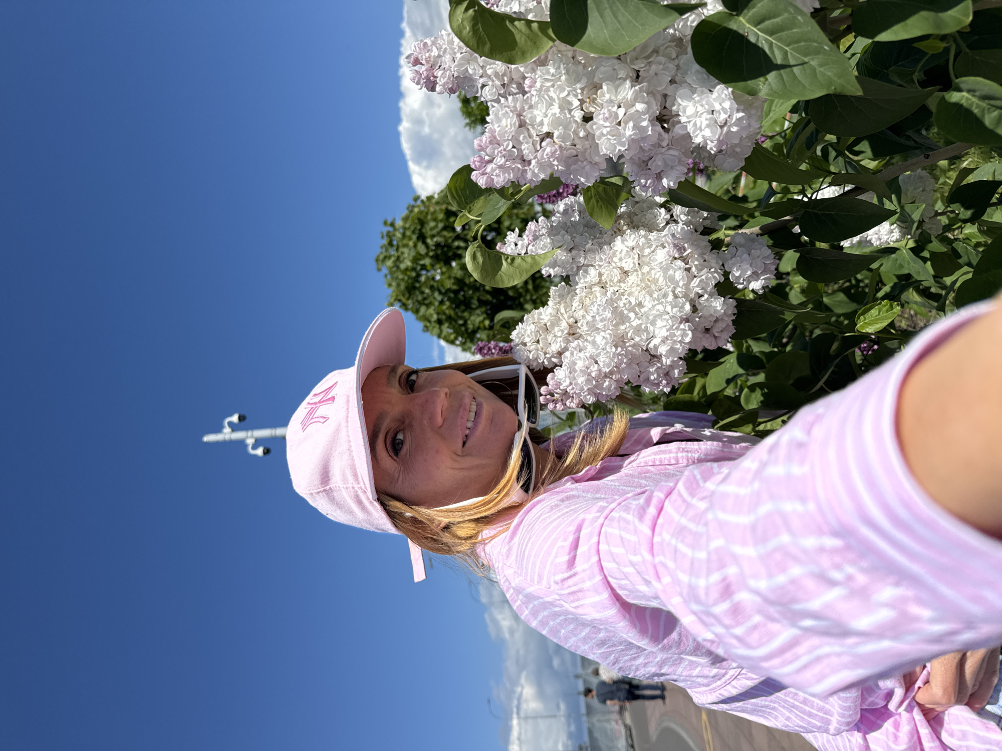 A woman is captured in a selfie-style shot, smiling directly at the camera, with a vibrant lilac bush in bloom next to her. She has blonde hair and is wearing a pink baseball cap with a white, stylized "ML" or similar logo on the front. Her attire includes a long-sleeved, light pink shirt with thin white stripes. White sunglasses are visible, pushed up on her head, framing her face.
The main background element is a dense lilac bush, filled with clusters of white and delicate light purple flowers and lush green leaves, occupying the right side of the frame and extending into the top right corner. The sky above is a clear, bright blue with a few scattered white clouds. In the mid-ground on the left, a tall, slender pole equipped with what appears to be multiple white cameras or light fixtures stands against the blue sky. The lower portion of the image shows hints of a paved area or road, possibly with a reflective surface and distant, blurred figures, suggesting an outdoor, public setting.
The scene takes place on a sunny day, indicated by the bright lighting and clear sky. The woman's cheerful expression conveys a positive mood. Based on the provided context, the image was taken in Saint Petersburg, Russia, capturing a moment of enjoyment amidst blooming nature.