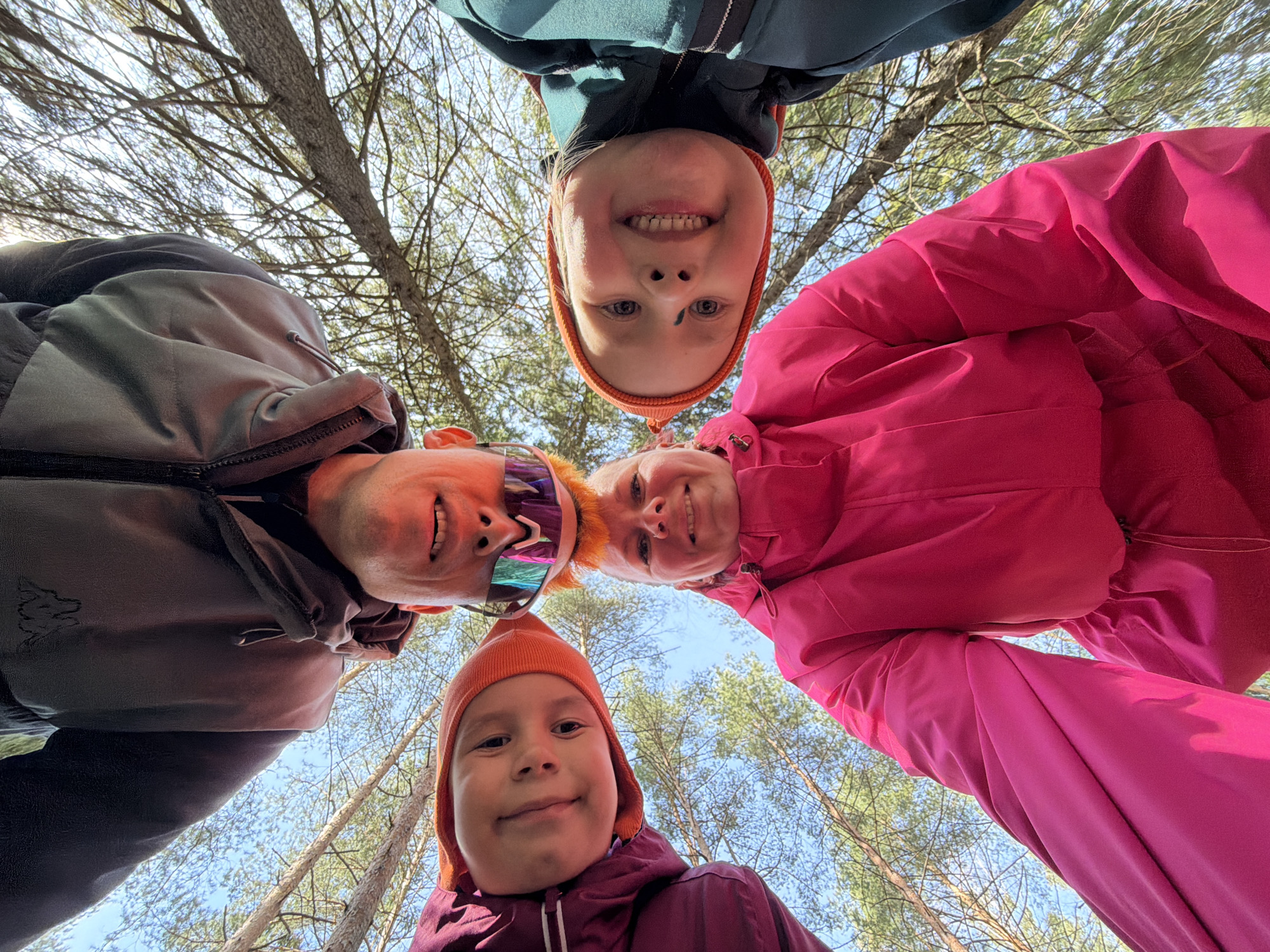 An upward-facing, low-angle group selfie captures four individuals—two adults and two children—looking directly down into the camera. The subjects are arranged in a circular formation, with their faces close to the lens. The background reveals a bright, clear blue sky punctuated by numerous bare tree branches and some green foliage, indicative of a forest or wooded setting in Chaikovsky, Russia.
The adult male, positioned on the left, wears a dark grey jacket with a faint animal logo, an orange beanie, and striking reflective sports sunglasses, smiling broadly. To his right, an adult female in a vibrant pink jacket and with short, light hair also smiles. Between them, a child in a teal jacket and an orange beanie beams, revealing a missing front tooth. At the bottom, another child, clad in a burgundy jacket and orange beanie, looks down with a gentle smile. The heavy jackets and beanies suggest cool temperatures, despite the bright, sunny daytime weather. The overall mood is cheerful, depicting a family or group enjoying an outdoor activity. No legible text is visible in the image.