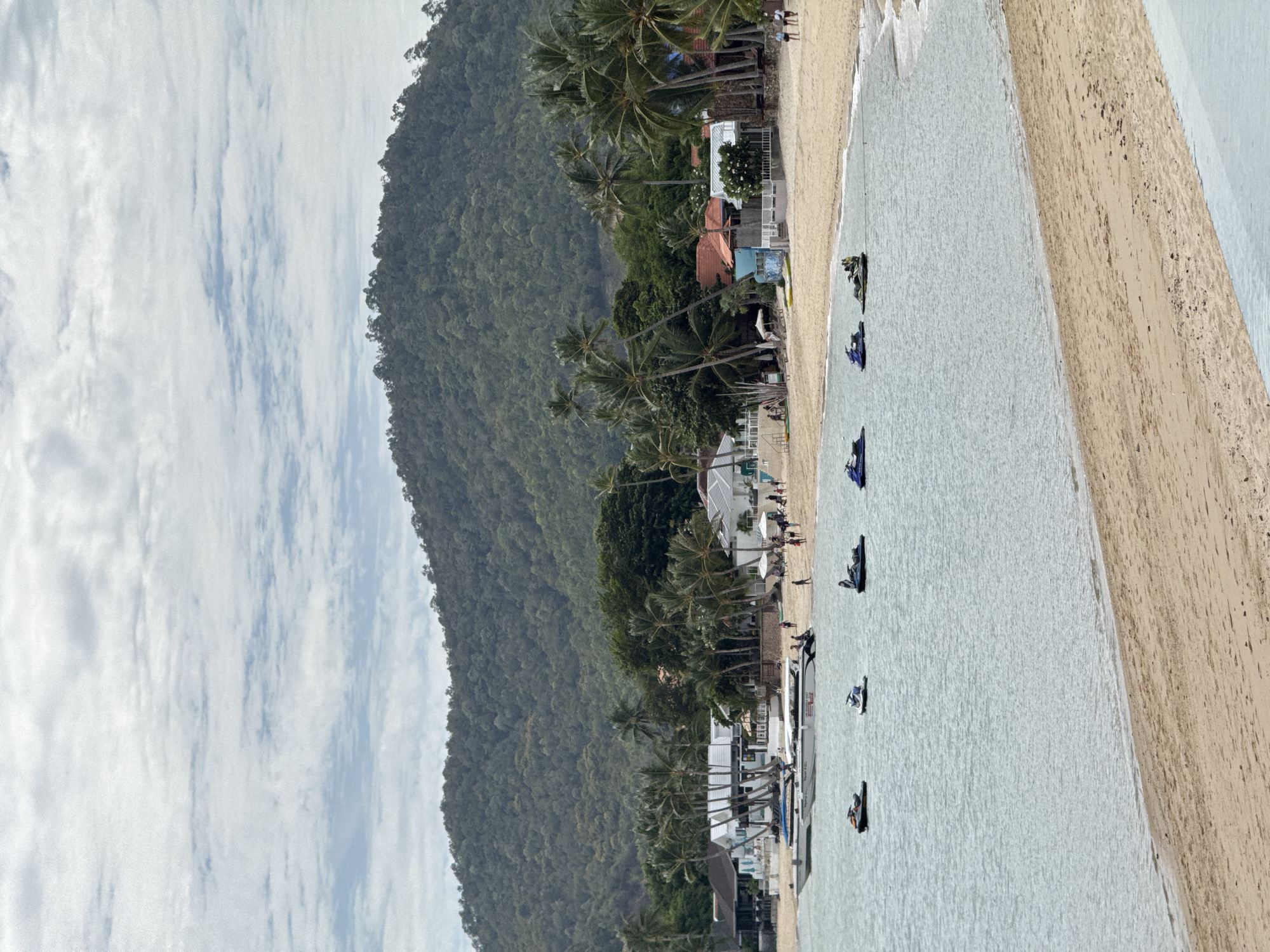 This is a wide, eye-level shot of a beach scene on Ko Samui District, Thailand. The image captures a sandy beach with calm, light blue water. Several jet skis are visible on the water, some with people riding them and others docked. A long, white boat with multiple passengers is anchored closer to the shore. The beach is lined with palm trees and several buildings, including what appear to be resorts or restaurants with white walls and tiled roofs. In the background, a lush green mountain covered in trees rises up, meeting a cloudy sky. There are small groups of people walking along the beach and near the buildings. The overall scene depicts a tropical vacation destination.