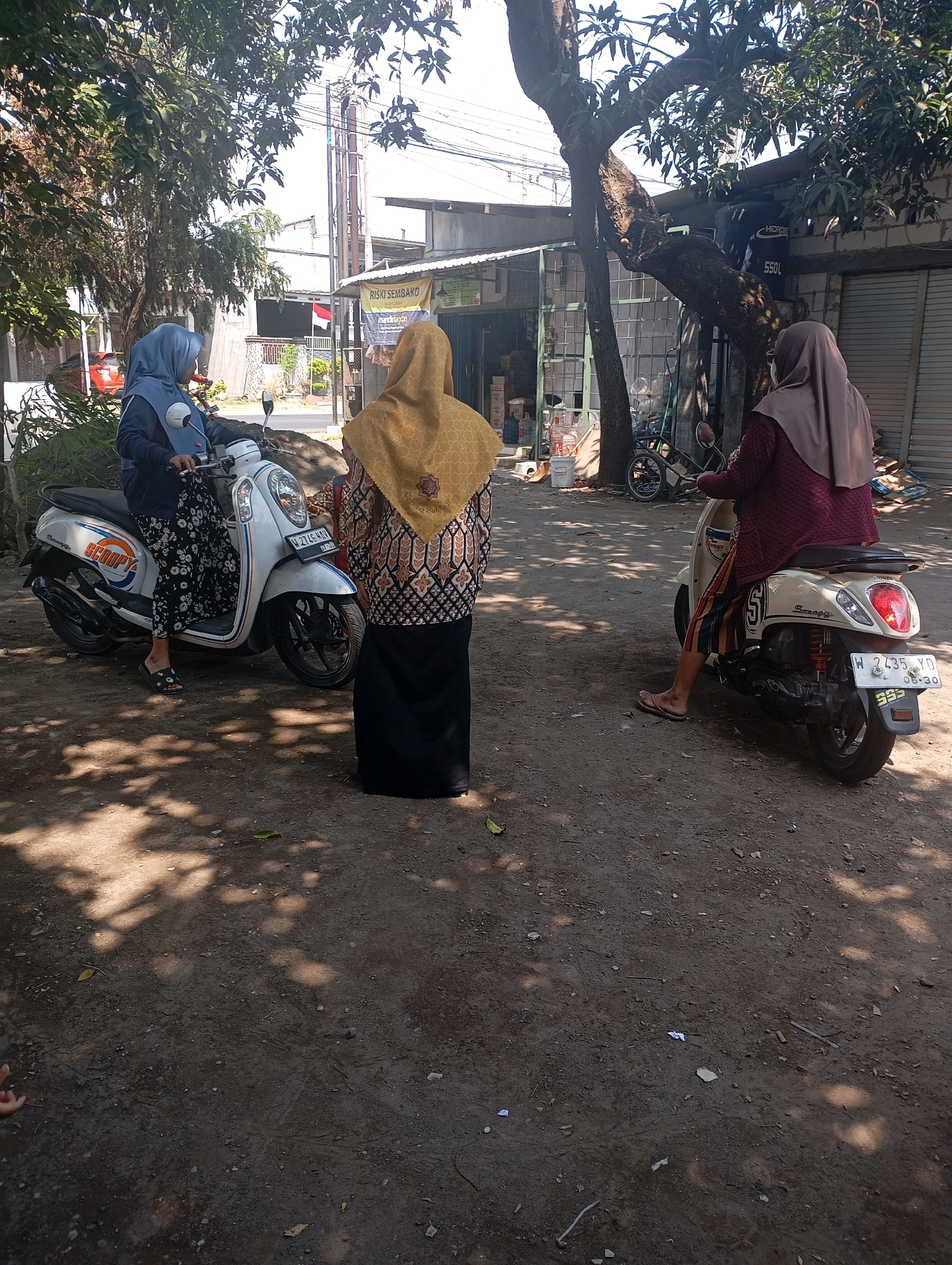 In Ketegan, Indonesia, an outdoor scene unfolds with three individuals and two motorcycles under the dappled shade of a large tree. On the left, a person wearing a blue hijab and a dark top with a floral skirt is seated on a white Honda Scoopy motorcycle, facing left. To the right of the first motorcycle, another individual in a yellow hijab and a patterned top with a black skirt stands facing away from the camera, their back to the viewer. Further to the right, a third person, seen from behind, is seated on a cream-colored Honda Scoopy motorcycle, wearing a light brown hijab and a maroon top with a striped skirt. The ground is a mixture of dirt and gravel, with scattered leaves and debris. In the background, a building with a corrugated metal roof and brick walls is visible, along with a sign that reads "RISIKI SEMBAKO." The overall atmosphere suggests a casual, everyday moment in a semi-urban environment.