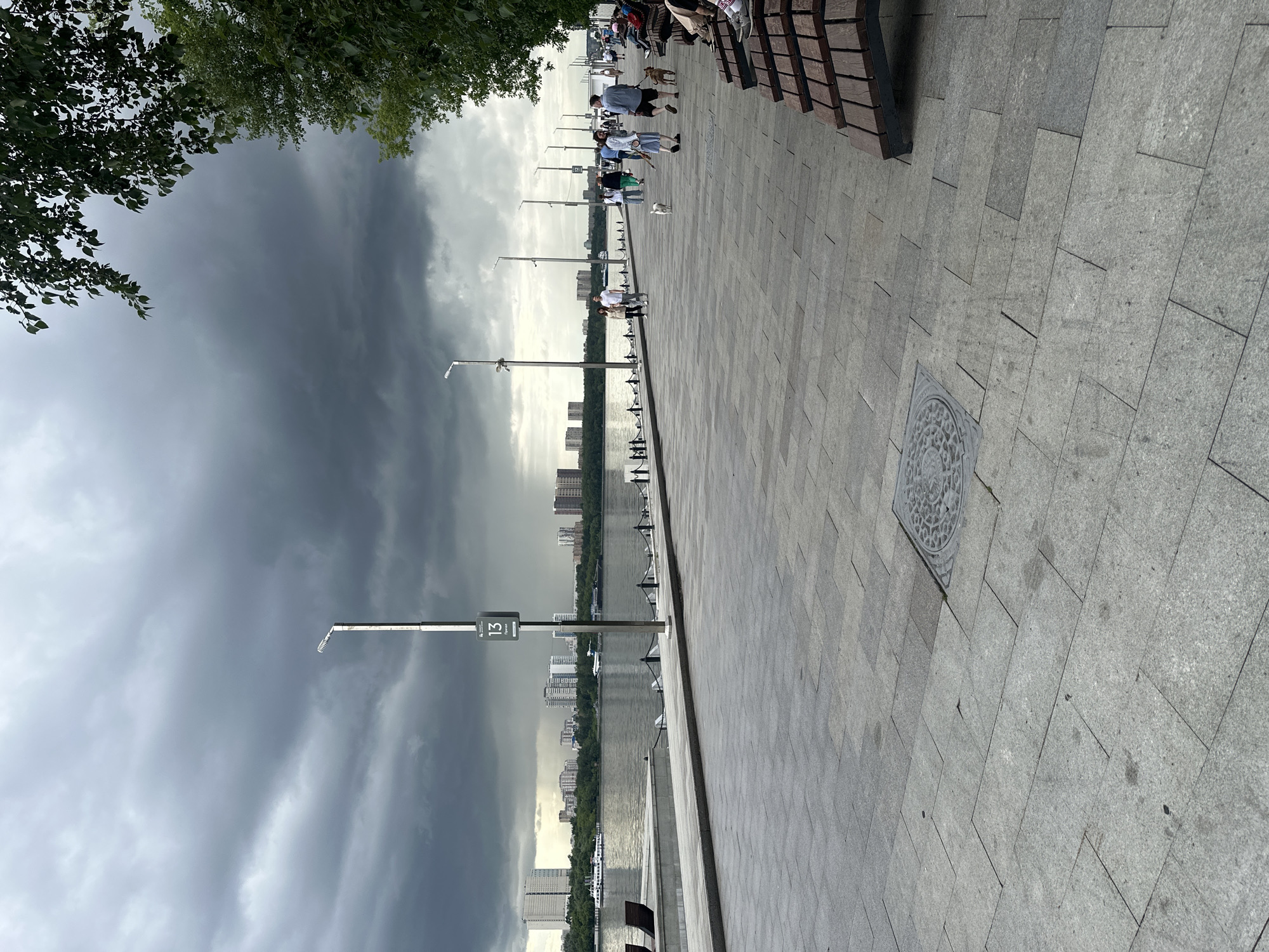 A wide shot shows a waterfront promenade with people walking and sitting on benches. The promenade is paved with large, grey tiles. To the left, a body of water is visible, with a cityscape in the distance. Dark, stormy clouds dominate the sky on the left side of the frame, contrasting with brighter skies on the right. Several lampposts are placed along the promenade. Some people are walking their dogs, while others are strolling casually. Benches are arranged along the edge of the promenade, with some people seated on them. A manhole cover with a circular pattern is visible on the pavement. A sign with the number "13" and some text is mounted on a lamppost. The overall mood appears somewhat somber due to the impending storm, yet there is a sense of everyday life continuing. The time of day is difficult to determine precisely due to the weather conditions, but the lighting suggests it could be afternoon.