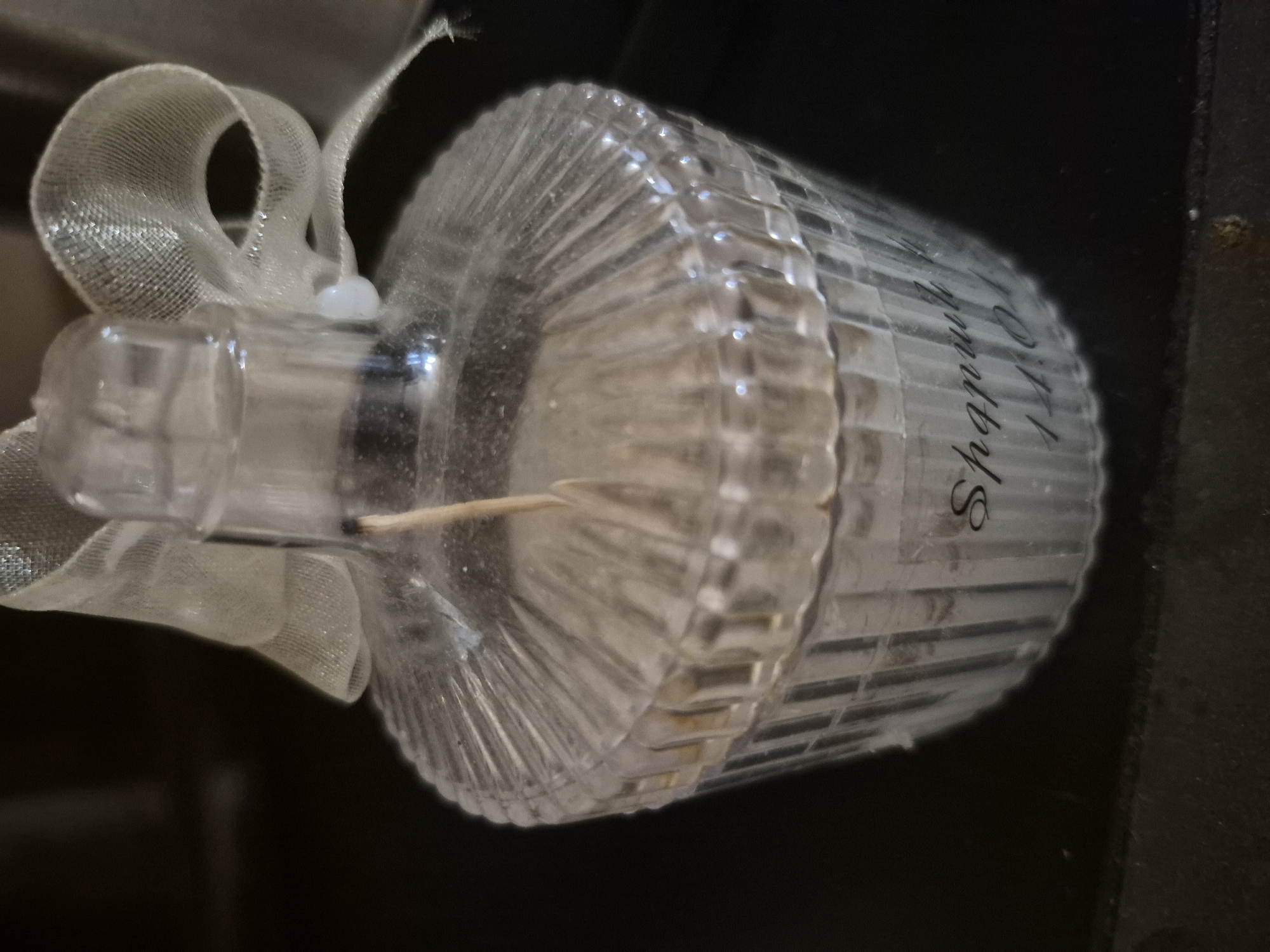 The media file presents a close-up, horizontally oriented image of a decorative clear container, likely glass or plastic, against a dark, indistinct background.
**Subjects:** The primary subject is a fluted cylindrical container, possibly a scented candle or diffuser. It is adorned with a sheer, light-colored ribbon and contains a small wooden stick.
**Objects Details:** The container has a narrow neck and a wider body characterized by prominent vertical ridges or fluting. Its transparent material reflects light, and some dust or particles are visible on its surface. Inside the container, a lighter, opaque substance, possibly wax or scented liquid, can be observed. A delicate, sheer ribbon, likely organza, is tied around the container's neck, forming soft, flowing loops. A small, round, white bead secures the ribbon knot. A thin, light brown wooden stick is inserted into the container's opening, suggestive of a diffuser reed or candle wick.
**Activity or Event:** No activity or event is taking place; the objects are static and presented as a still life.
**Notable Details:** The image appears to be taken indoors under artificial lighting, as indicated by the dark and featureless background. There are no visual cues to determine the time of day, weather, or specific location within Yerevan, Armenia, other than the provided context. The overall impression is that of a handcrafted or decorative home fragrance item.
**Visible Text:** Text is visible on the side of the fluted container, written vertically in a decorative, cursive script. It clearly reads "D.I.Y. Aromas" followed by another word that appears to be "Shopsy" or "Shopsy's".