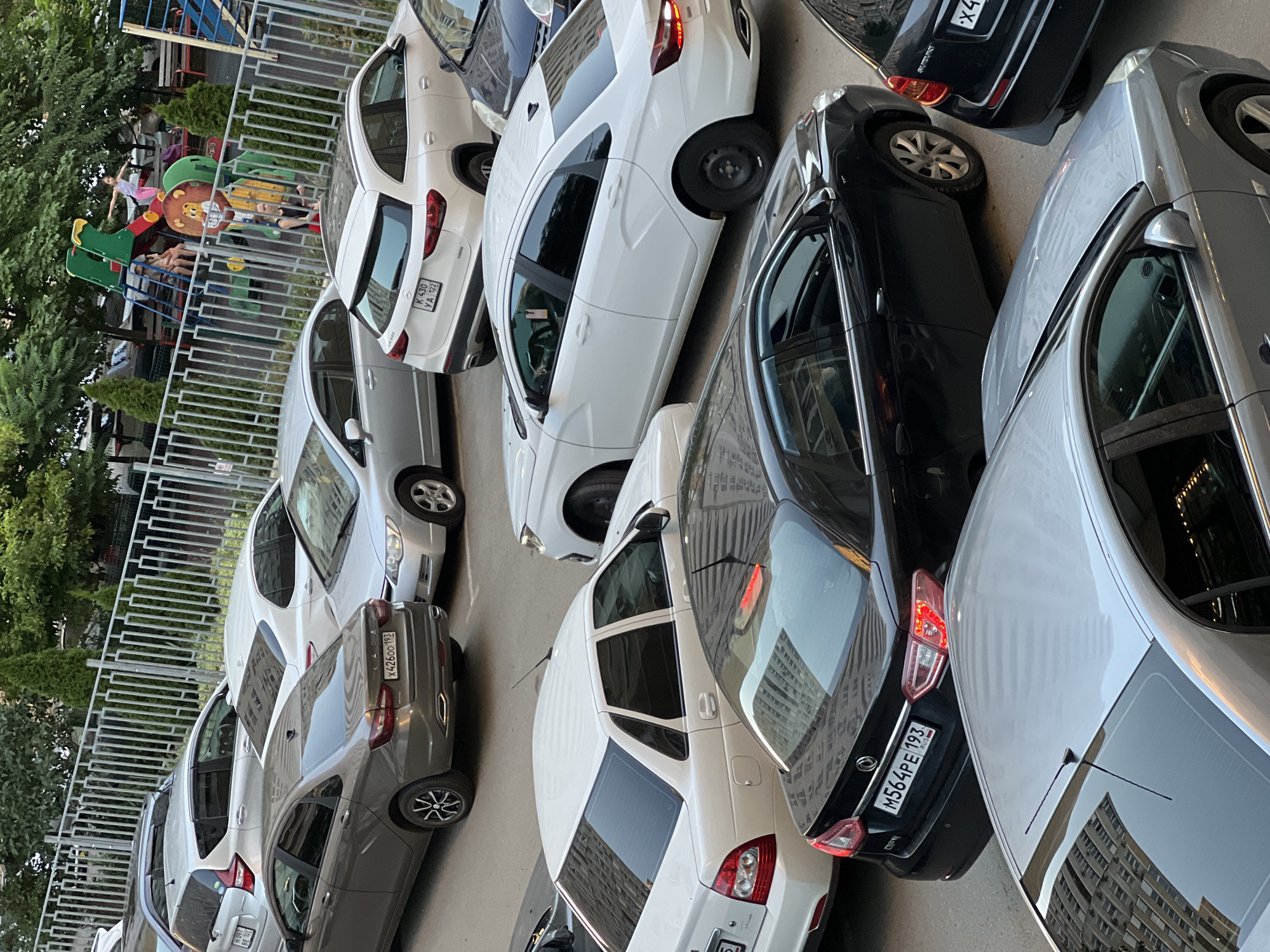 The image depicts an outdoor parking lot in Krasnodar, Russia, adjacent to a children's playground. Several cars are parked, including a silver sedan with license plate "X426OO 193", a white sedan with license plate "K 430 YA 122", a dark sedan with license plate "M564PE 193 RUS", and other vehicles that are partially obscured. Children are visible on the playground, with one child on a slide and others sitting. Trees and a fence line the perimeter of the playground. The overall scene suggests a residential area with recreational facilities.