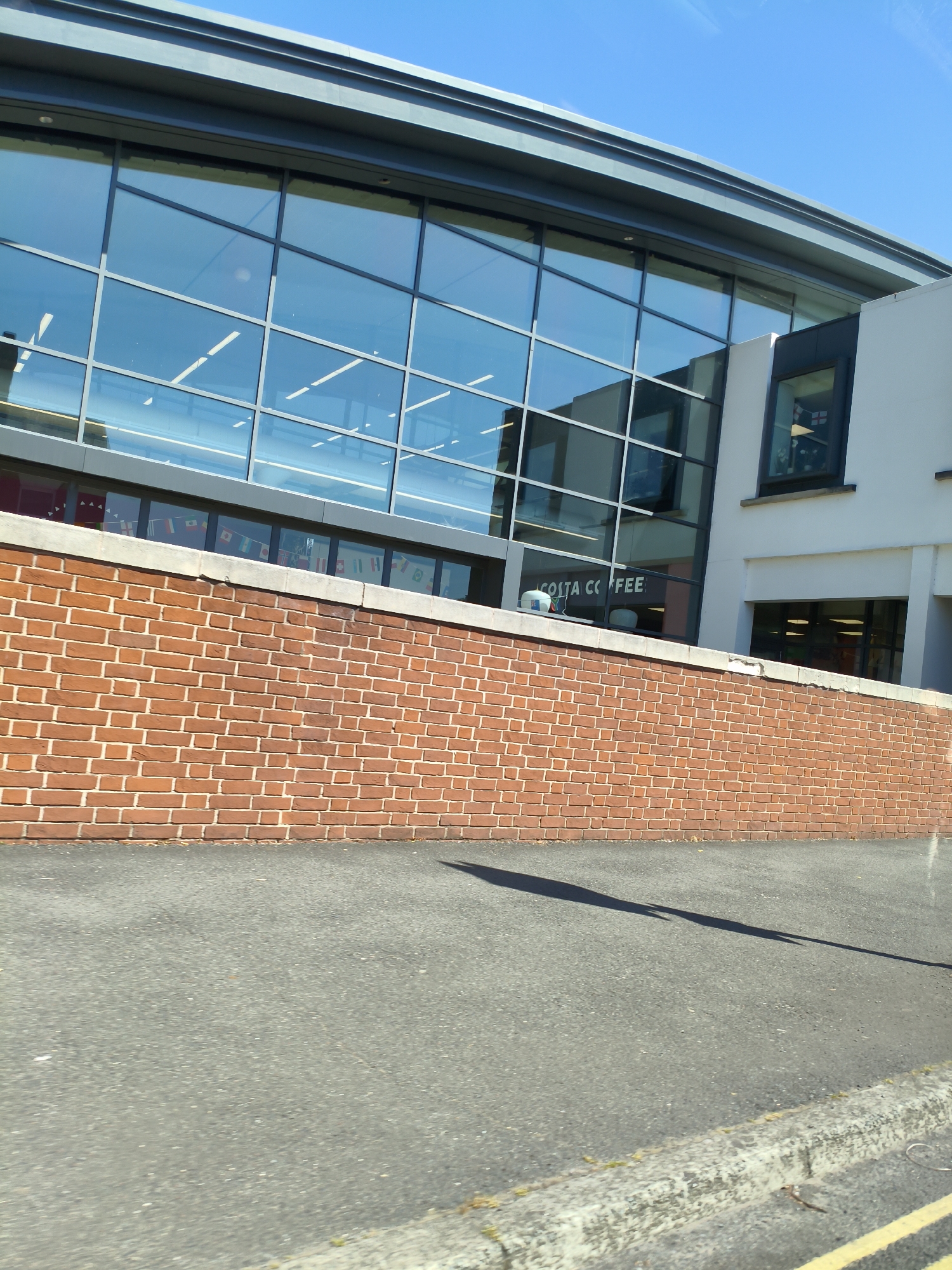 The image shows a modern building with a large glass facade, reflecting the blue sky and some interior lights. Below the glass facade is a brick wall, and in front of that is an asphalt road. The building appears to be a commercial establishment, as indicated by the "COSTA COFFEE" sign visible through the glass. A small window on the right side of the building shows a flag, possibly the Union Jack. The scene is set on a sunny day. The location is Ludlow, United Kingdom.