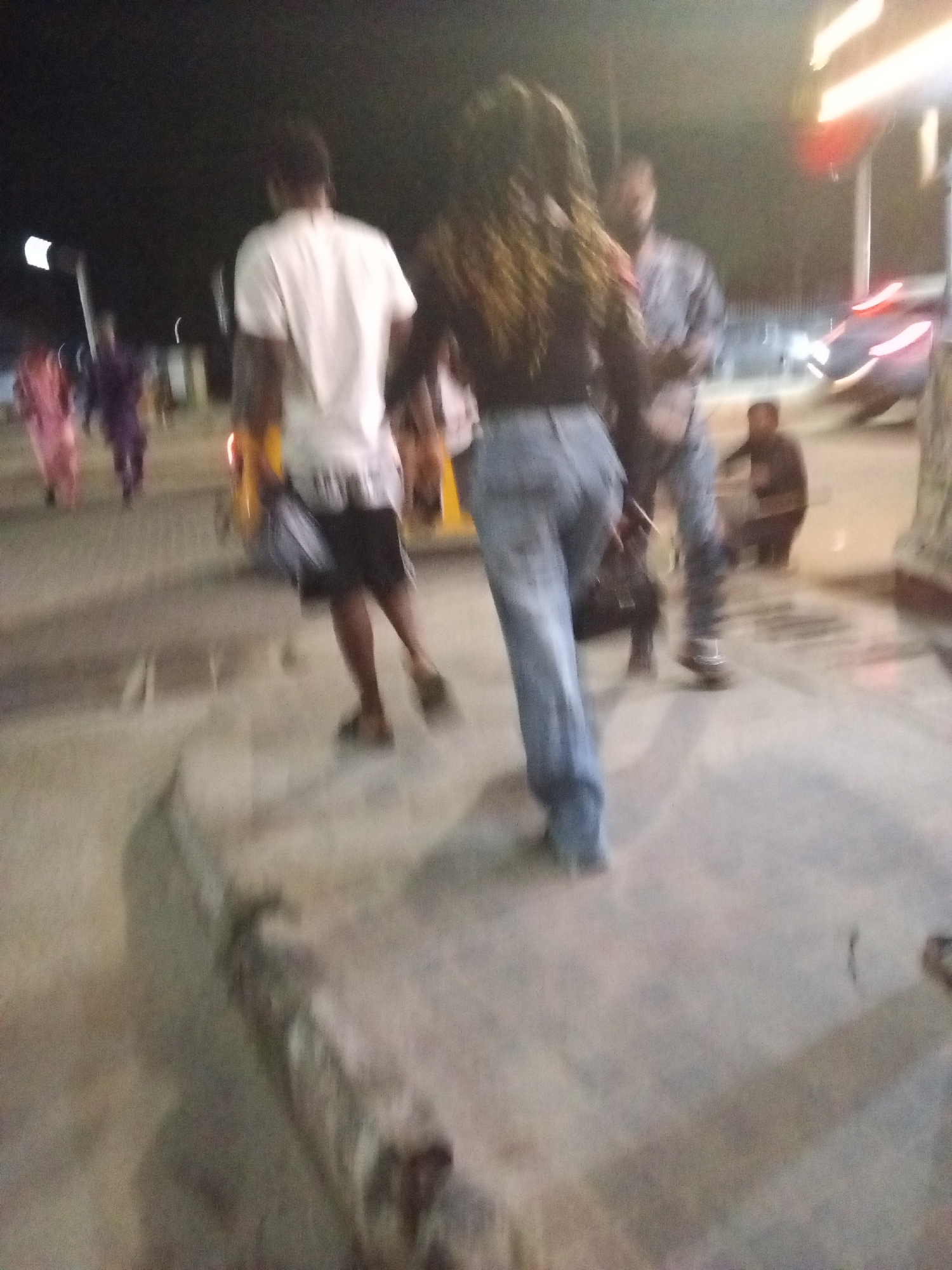 The image shows a street scene at night in Ikorodu, Nigeria. The location appears to be a busy area with pedestrians and vehicles.
Several people are walking on the sidewalk or street, including a man wearing a white t-shirt and dark shorts, a woman with long, wavy hair wearing jeans and a dark top, and another man wearing a light-colored long-sleeve shirt. There are other people walking in the background as well.
In the background, there is a car parked, street lights, and what appears to be a building with signage. The overall lighting is dim, with artificial lights illuminating parts of the scene.