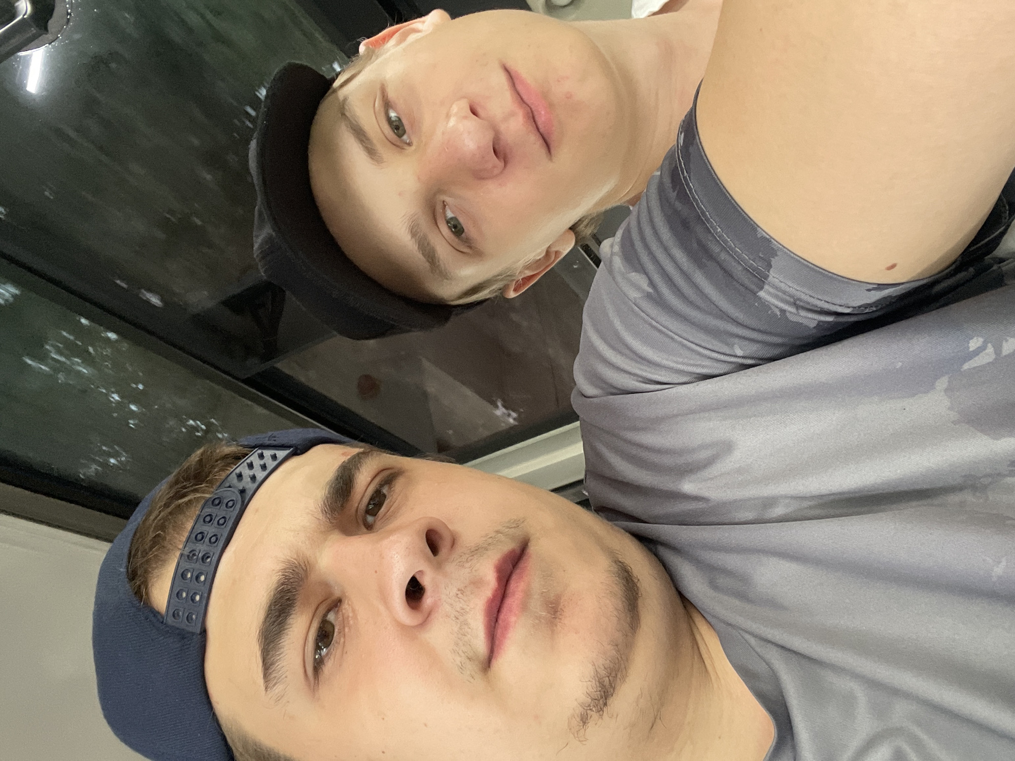 In a close-up selfie taken outdoors in Oskemen, Kazakhstan, two young men are captured. The man on the left, wearing a navy blue baseball cap and a grey t-shirt, is positioned closer to the camera. He has brown eyes, dark eyebrows, and light stubble on his chin and upper lip. The man on the right, wearing a black baseball cap, has light green eyes and appears to be slightly further away. Both men are looking towards the camera, suggesting they are posing for the photo. The background shows a dark, reflective surface, possibly a window or a car roof, with some blurred foliage visible. The lighting suggests it might be daytime.
