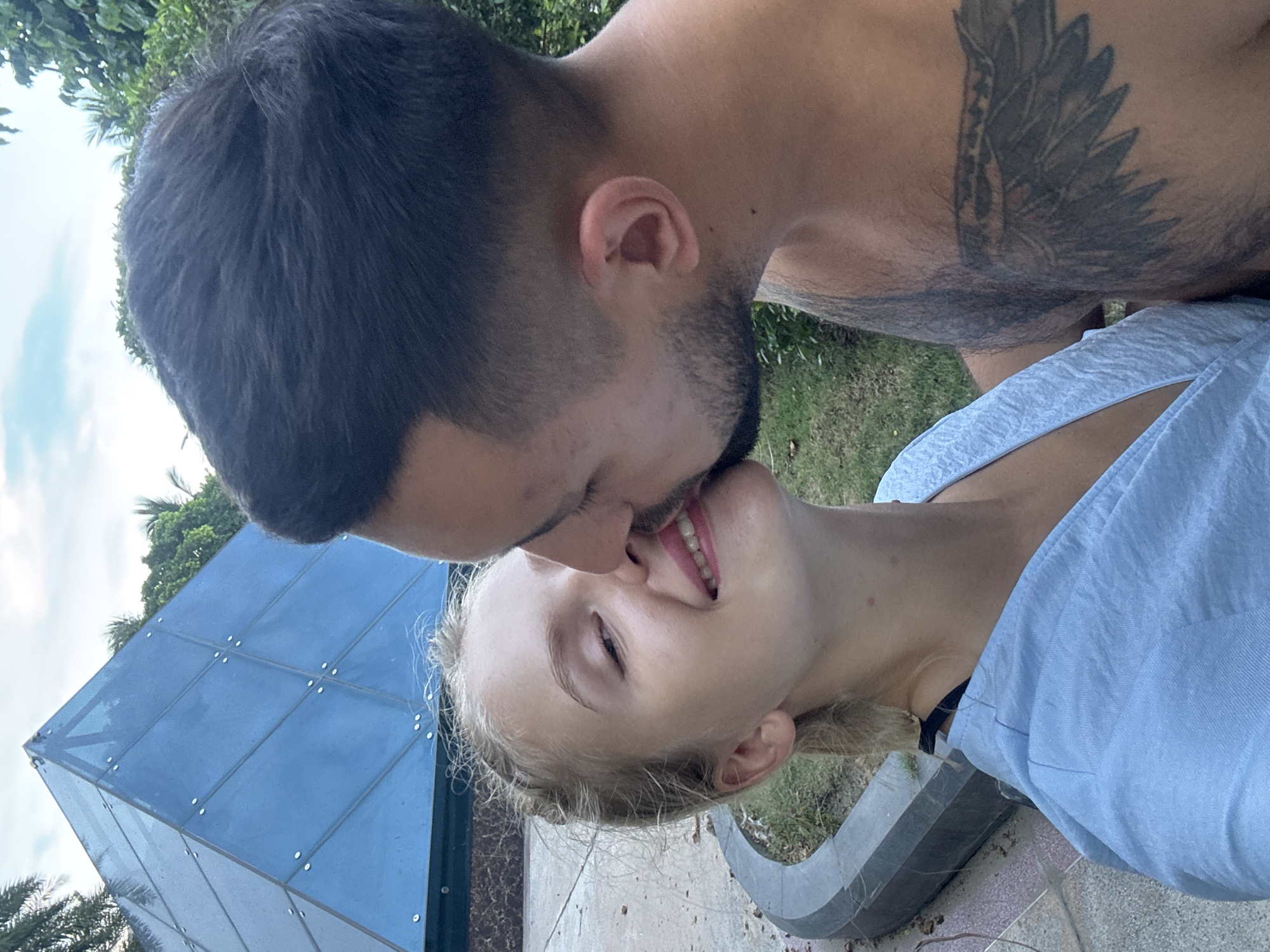 A young Caucasian couple is captured in a close-up outdoor photograph, sharing a kiss. The woman, with blonde hair styled in braids, smiles with her eyes partially closed as the man, with short dark hair and stubble, leans in to kiss her. The man has a tattoo of a wing on his chest. In the background, a blue glass pyramid structure is visible, along with lush green foliage and a cloudy sky. The scene appears to be a public area, possibly a park or a resort, with a paved walkway and a curved concrete barrier in the foreground. The environment suggests a tropical location, consistent with Nha Trang Ward, Vietnam.