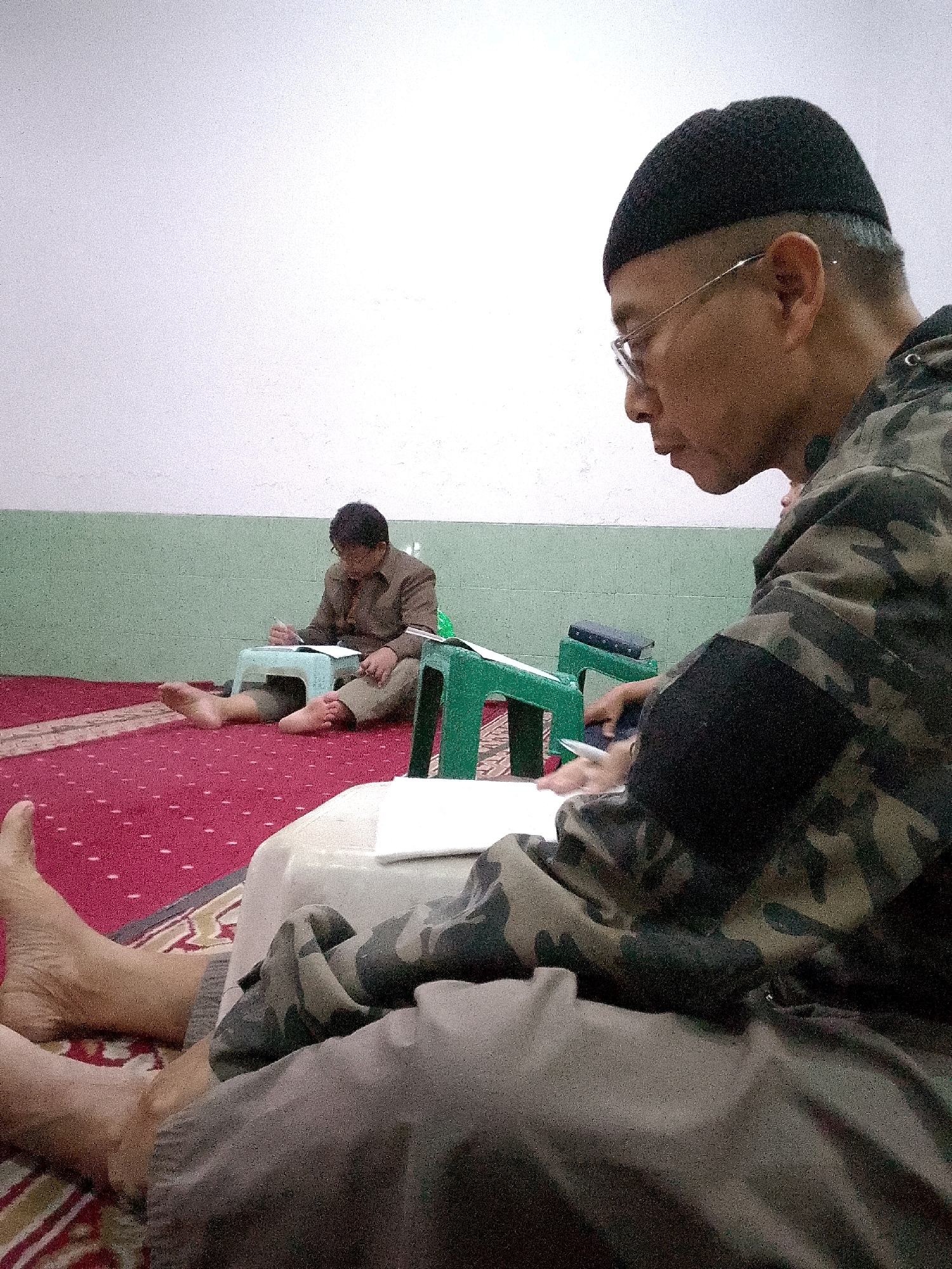 In a room with a white and green speckled wall, two individuals are seated on a red carpet patterned with small white dots. In the foreground, a person wearing a black beanie, glasses, and a camouflage jacket is looking down at a white piece of paper and appears to be writing. Their legs are crossed, and they are barefoot. In the background, a younger individual wearing a brown shirt and pants is also seated on the carpet, looking down at a book placed on a small, light blue stool. This person is also barefoot and holding a pen. Two green plastic stools are present in the room, one of which is being used to support the book in the background. The overall scene suggests a quiet study or religious gathering in Gunungmasigit, Indonesia.