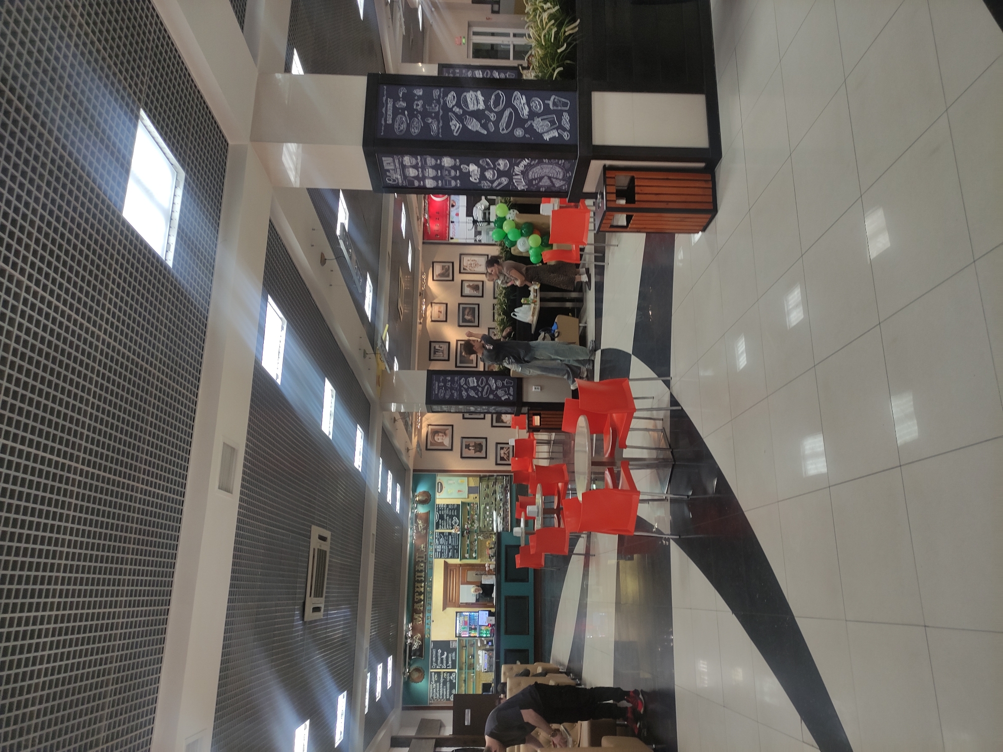 The image shows the interior of a cafe or restaurant in Pavlodar, Kazakhstan. A main structural column with a menu board stands in the space. Several people are gathered near tables and chairs, some of whom are interacting, while others are standing or walking. A service counter with a menu board is visible in the background, indicating the point of sale. Rows of black and white square floor tiles create a pattern across the room. A grid-patterned ceiling with light fixtures and ventilation systems is overhead.