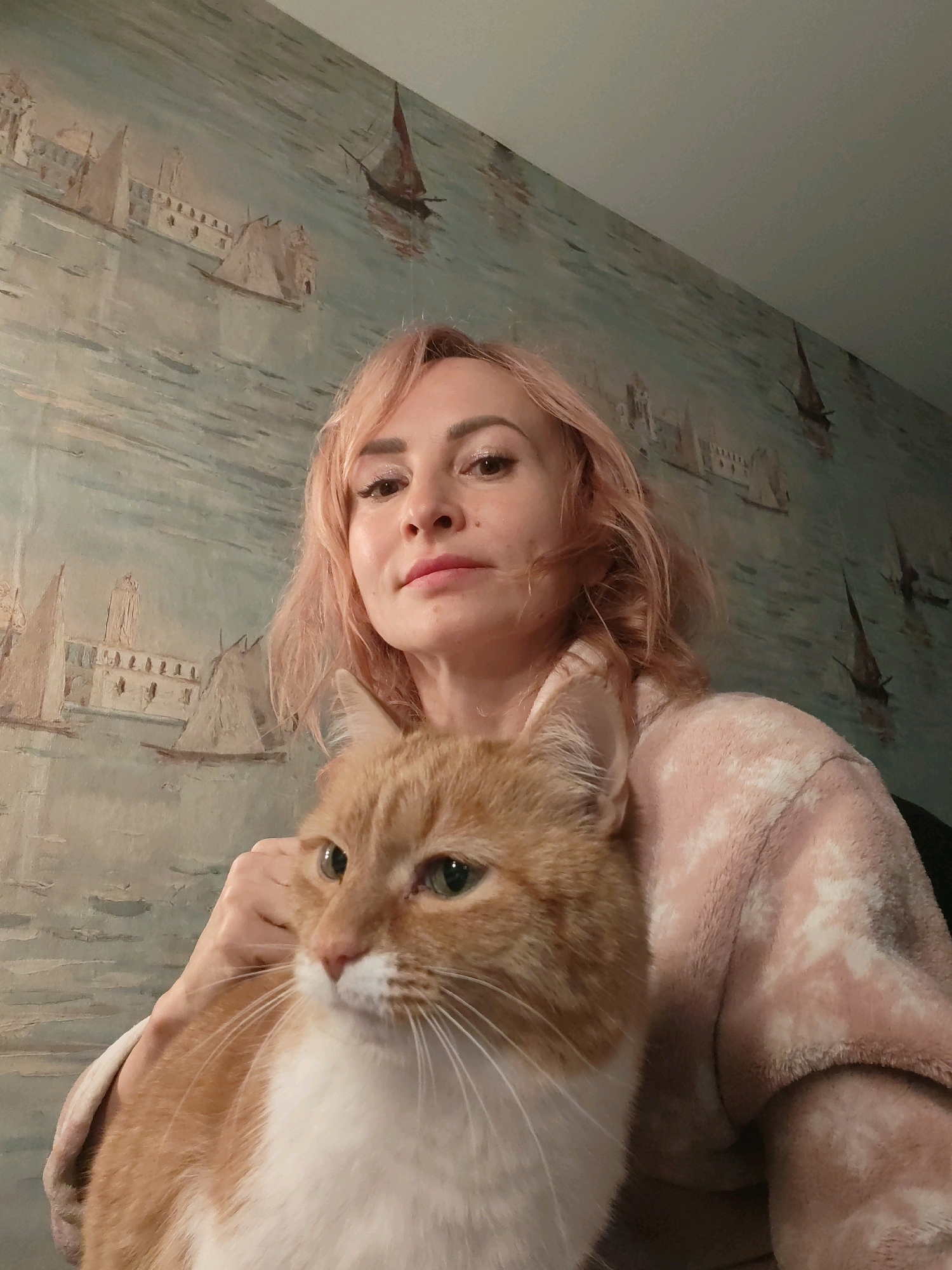 A woman with short, pink hair and wearing a light pink bathrobe is holding an orange and white cat. The cat is in the foreground, looking to the left. The woman is in the background, looking towards the camera. The background is a wall with a mural depicting sailboats on a body of water, with buildings on the shore. The image appears to have been taken indoors. There is no visible indication of the city of Chaikovsky, Russia in the image.