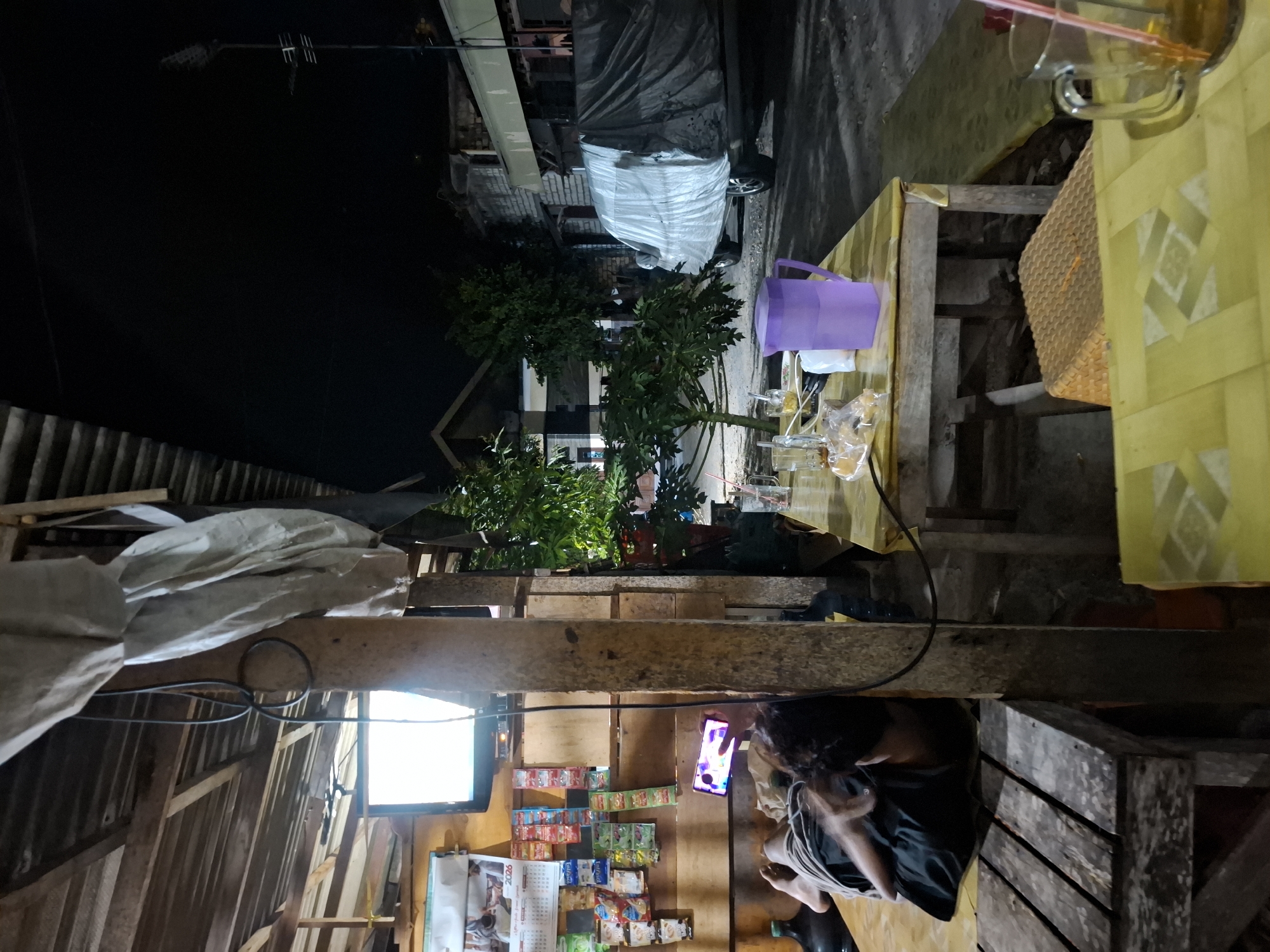 A dimly lit outdoor seating area in Bulubrangsi, Indonesia features wooden tables and benches, some covered with yellow mats. A person is lying on a mat, looking at a phone, with a large water jug nearby. On a table, there is a purple pitcher, drinks in glasses, and some snacks. In the background, a vehicle is covered with a tarp, and a television displays a bright screen. The scene appears to be a roadside eatery or rest stop.
