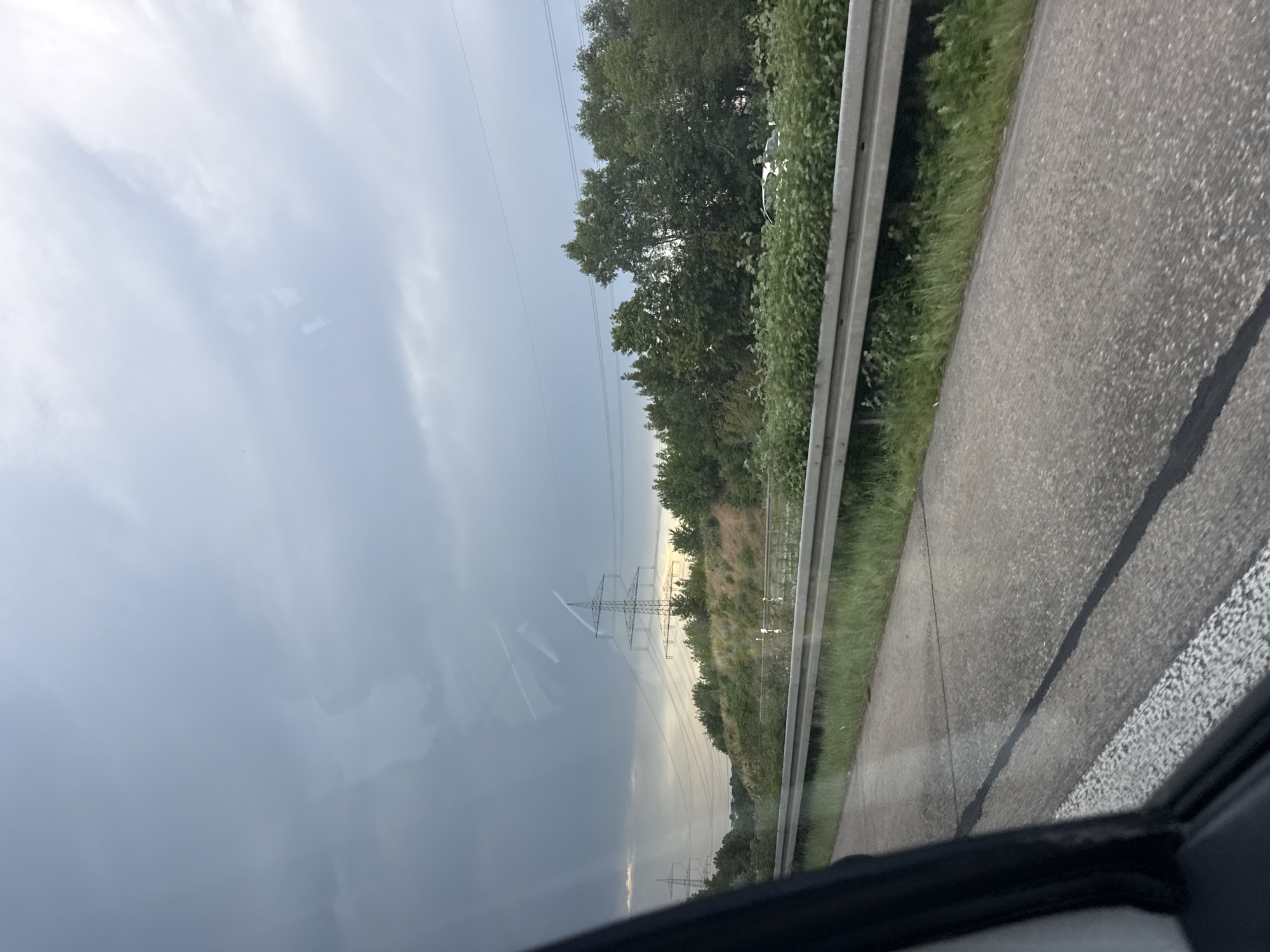 The image depicts a view from a vehicle traveling on a highway. The sky is overcast with dark clouds. In the background, there are several large power pylons with electrical wires. A line of trees and vegetation runs alongside the highway. On the right side of the highway, there is a white car visible amongst the greenery. The highway itself is paved and has a metal guardrail separating it from the roadside. The scene appears to be captured in the vicinity of Linden, Germany.