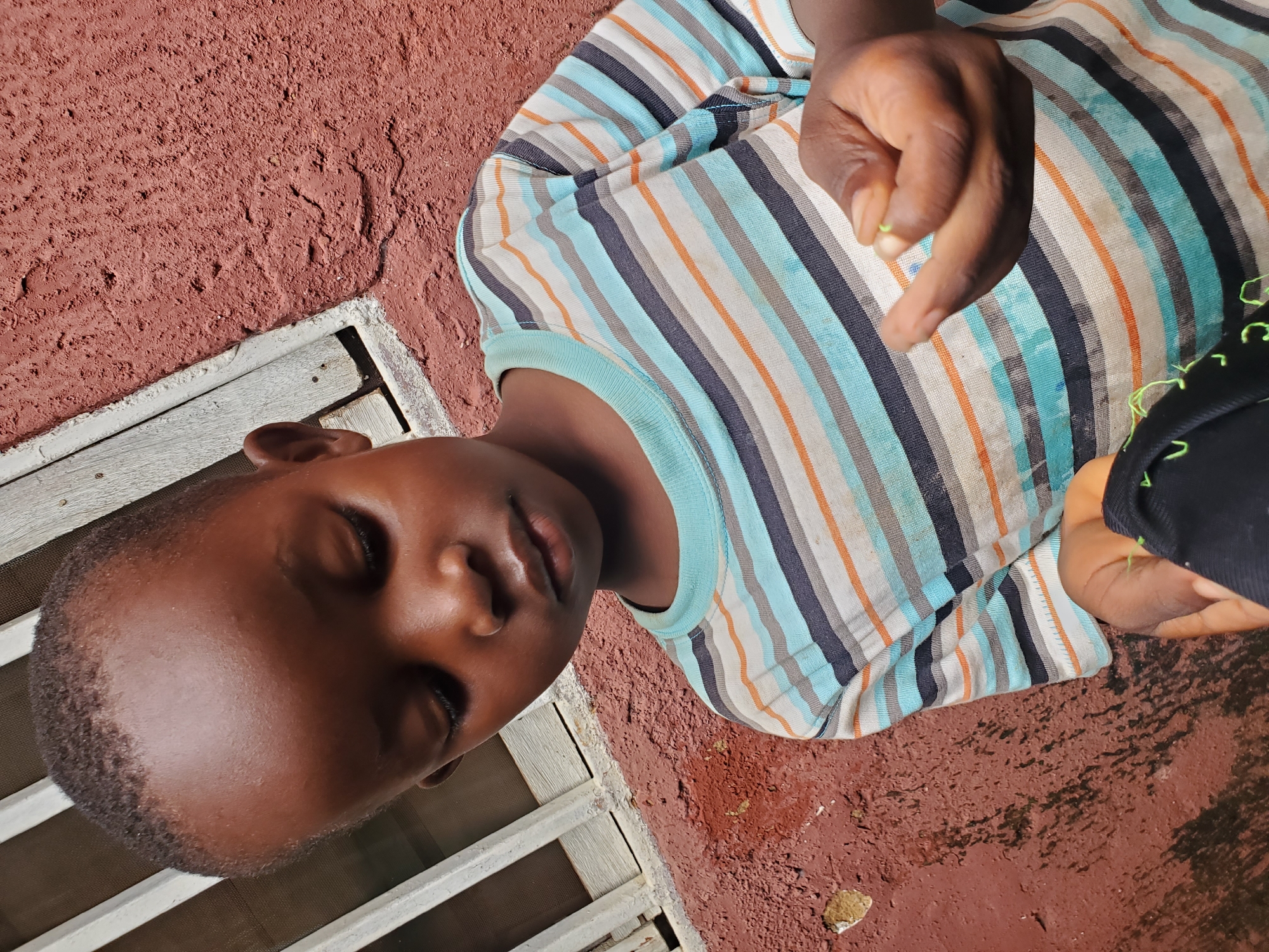 This is a close-up, sideways shot of a young boy lying down, with his eyes closed. He is wearing a striped t-shirt with horizontal stripes in blue, white, orange, and black. His dark skin is visible, and his hair is short and dark.
The boy is lying on a reddish-brown, textured surface that appears to be a wall or ground. To his left, there is a white wooden structure, possibly part of a window or grate. The lighting suggests it is daytime, with a shadow cast across the scene. A hand is visible in the upper right corner of the frame, with a small green object held between the fingers. The overall impression is one of a casual, outdoor setting. The location context of Jos, Nigeria, suggests this could be a typical scene in a residential area.