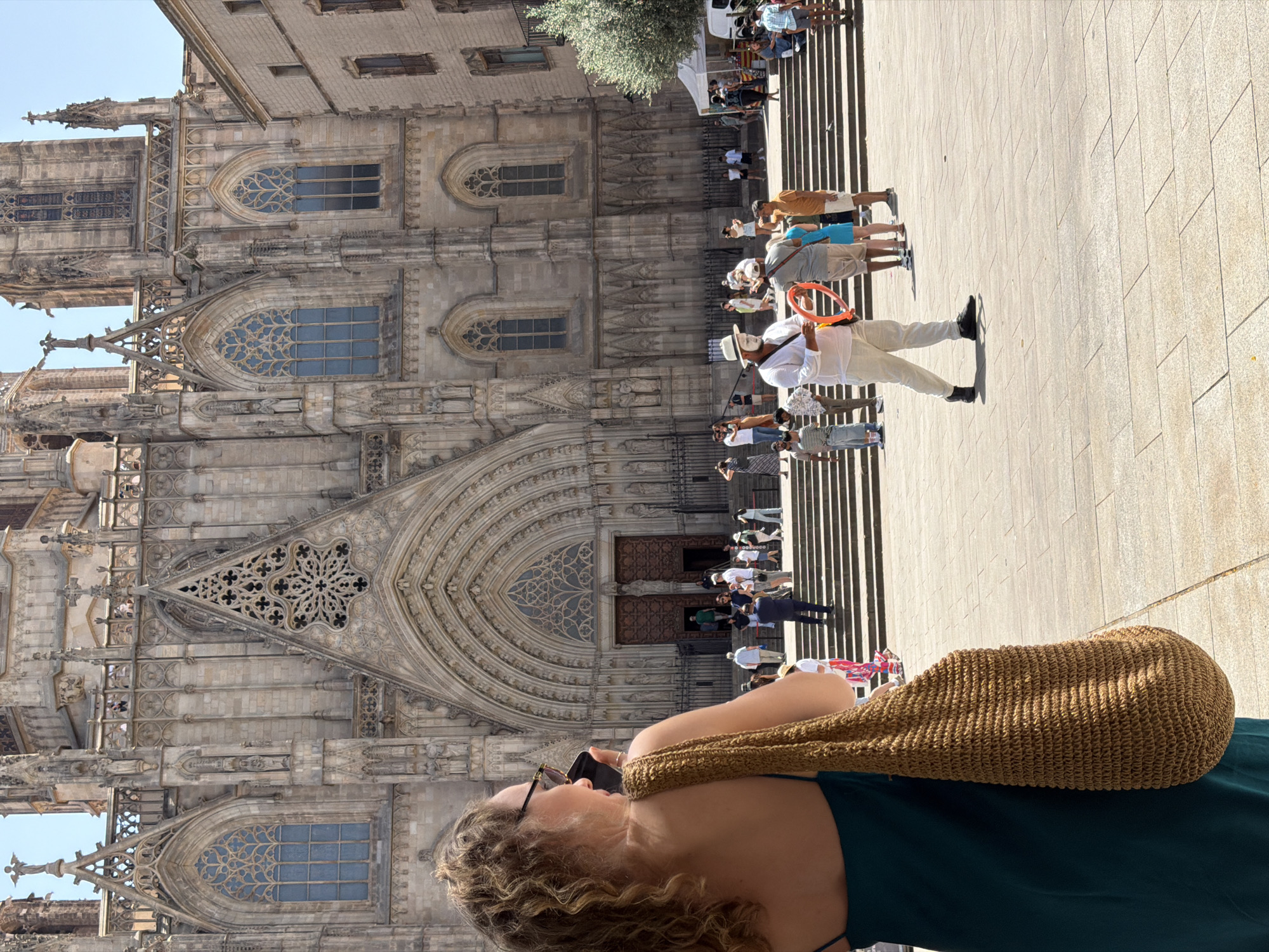 A woman with curly hair and sunglasses is standing in the foreground on the right, holding a smartphone in her right hand and a large woven bag over her left shoulder. She is facing away from the camera, looking towards a large Gothic cathedral. In the middle ground, a group of people are gathered on a wide set of stairs leading up to the cathedral entrance. A performer in a white outfit and white face paint is holding an orange ring in the center of the crowd, interacting with onlookers. The cathedral is made of stone and features ornate architectural details, including arched doorways and stained-glass windows. A white tent is visible on the far right, and trees are present in the background. The scene appears to be taking place in Barcelona, Spain, likely in front of the Barcelona Cathedral, given the architectural style. The ground is paved with large square tiles. The lighting suggests it is a bright, sunny day.