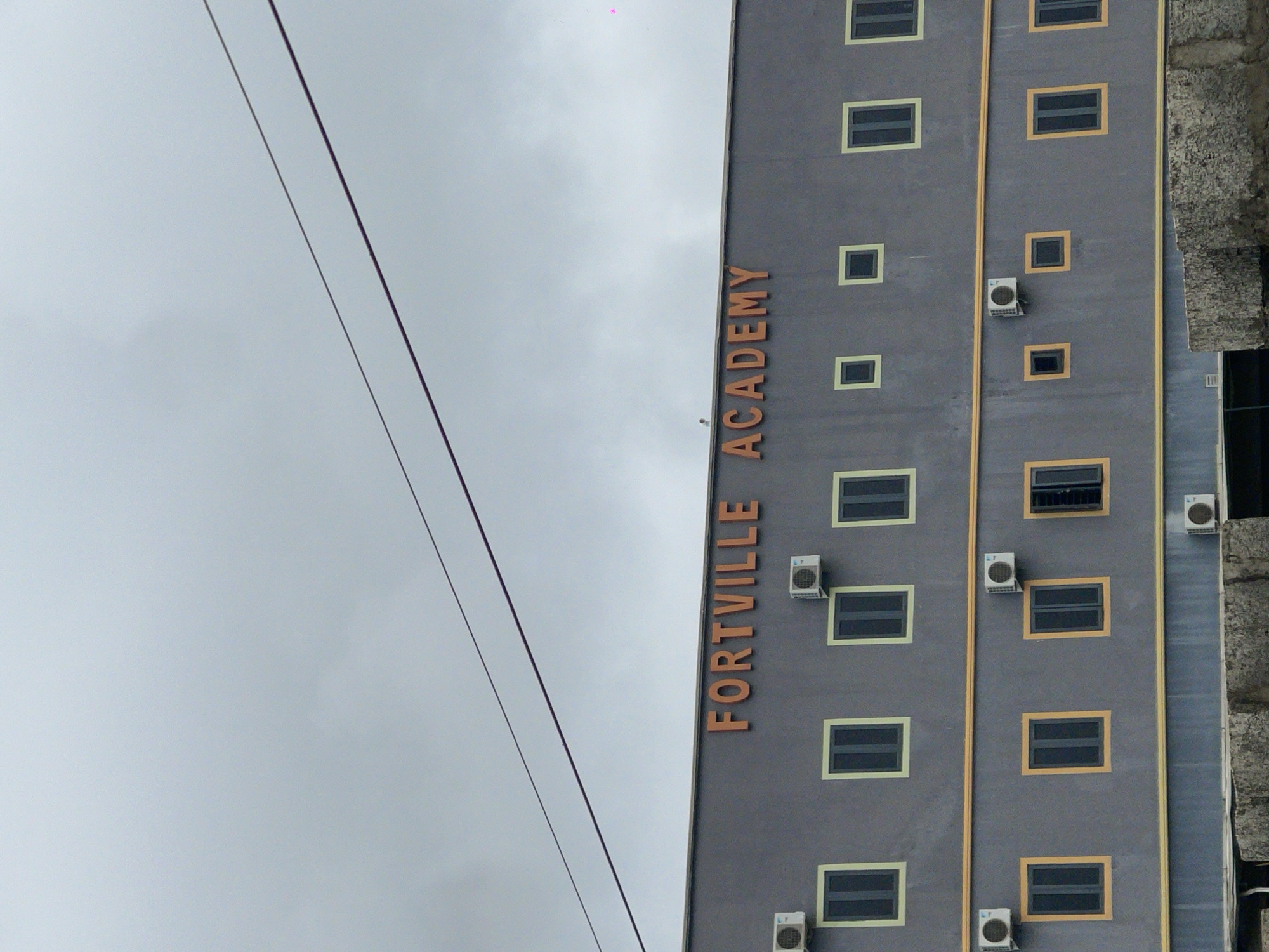 The image shows a multi-story building with a dark gray facade, identified by orange lettering as "FORTVILLE ACADEMY". The building has a series of windows, some with light yellow frames and others with dark frames. Several air conditioning units are mounted on the exterior walls. Two power lines run diagonally across the cloudy sky above the building. The context provided states this is in Abuja, Nigeria, though the image itself does not contain any explicit geographical markers. The scene appears to be an exterior shot of an educational institution.