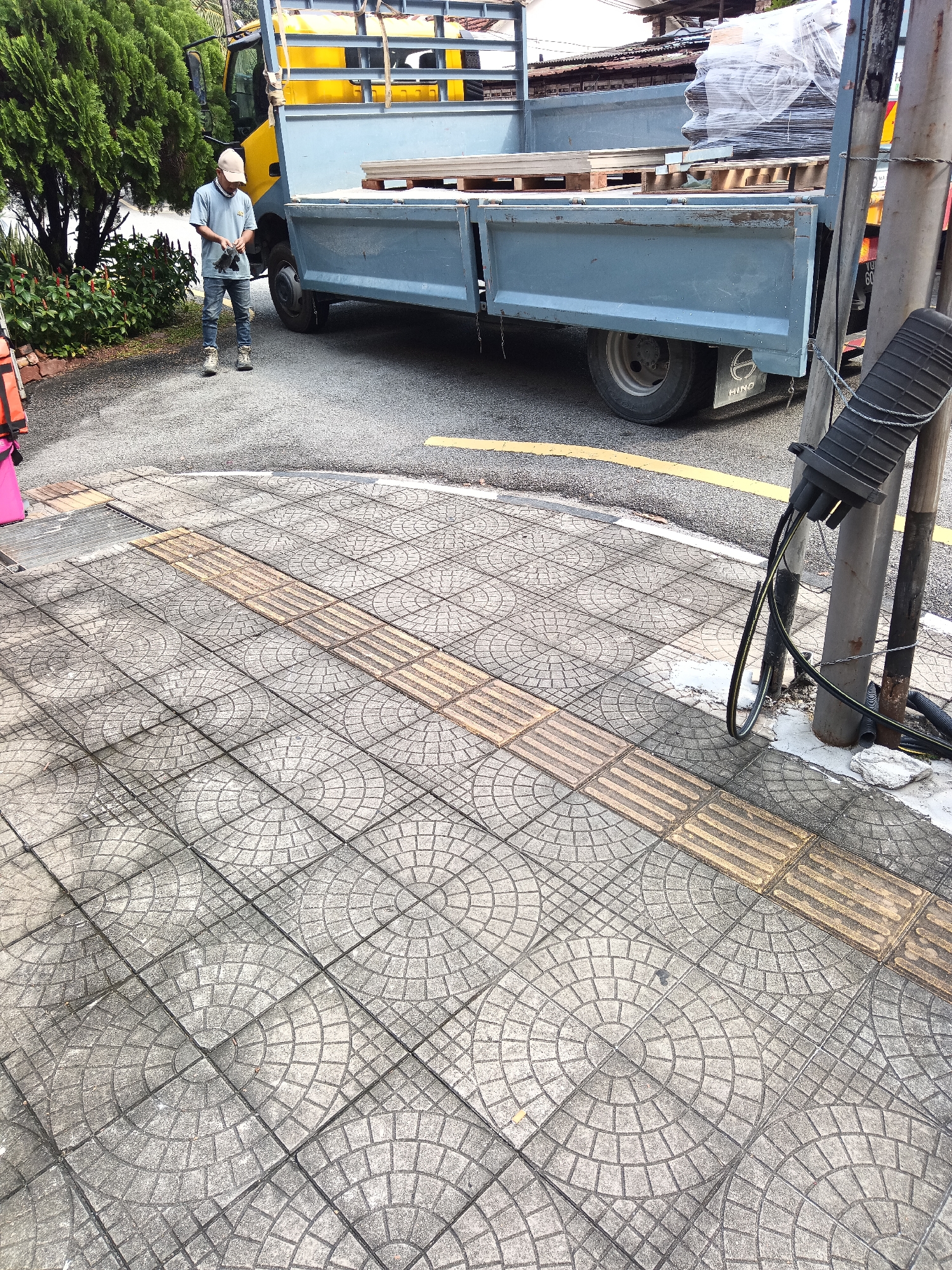 The image depicts a man wearing a light-colored cap, t-shirt, and jeans, standing on a sidewalk paved with circular patterned tiles. He is holding black gloves in his hands, seemingly preparing to put them on. Behind him, a blue truck is parked. The truck has a yellow cabin and a flatbed loaded with wooden pallets and materials wrapped in plastic. The sidewalk also features tactile paving, indicating a pedestrian pathway. In the background, lush green trees and foliage are visible, suggesting a potentially urban or suburban environment. A utility pole with cables attached is located to the right of the frame, near the sidewalk edge. The scene appears to be an outdoor setting, possibly in Kuala Lumpur, Malaysia, given the visual elements and common urban infrastructure found there.