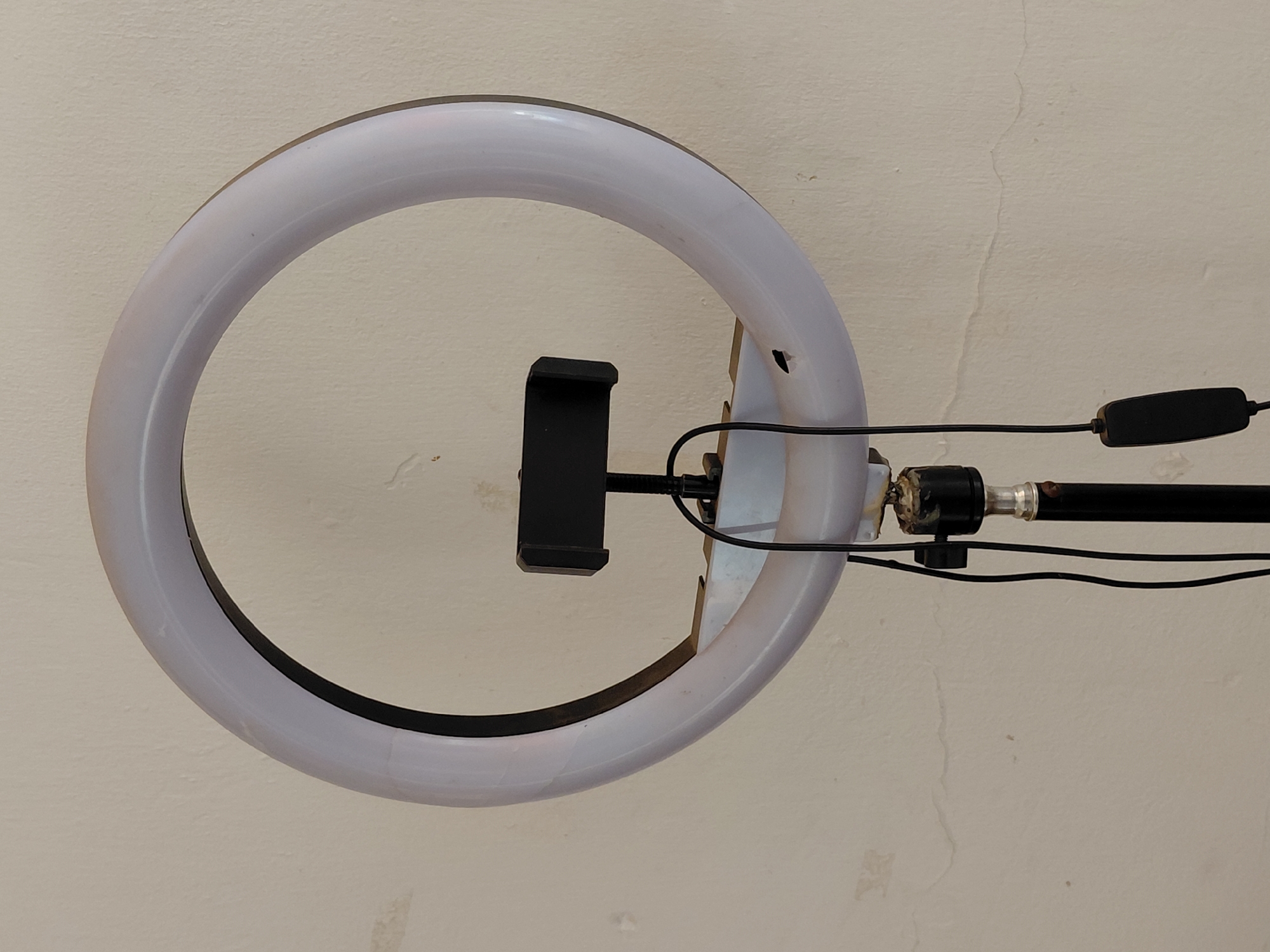 An indoor, eye-level shot captures a white, circular ring light with a phone holder mounted in its center, against a textured, light-colored wall. The ring light itself appears to be made of translucent white plastic, with a darker, possibly black, inner rim. A black, spring-loaded phone holder is positioned in the middle, ready to secure a smartphone for recording or streaming.
Black electrical cables emerge from the right side of the ring light, connecting to an inline control unit, also black, which is visible further along the cable. These wires run along a black pole or stand component that supports the light. The connection point between the ring light and its stand shows some wear and a brownish, possibly adhesive or repair material.
The background wall is a dominant feature, displaying an off-white or cream color with noticeable textural irregularities. Several fine, vertical cracks are visible on the wall's surface, particularly in the upper right and lower left sections, suggesting age or structural movement. There are also subtle areas of discoloration or staining on the wall. The overall impression is an interior setting, likely a room, with the ring light poised for use in content creation, such as video recording or photography. No people are visible, nor is any discernible text present in the image.