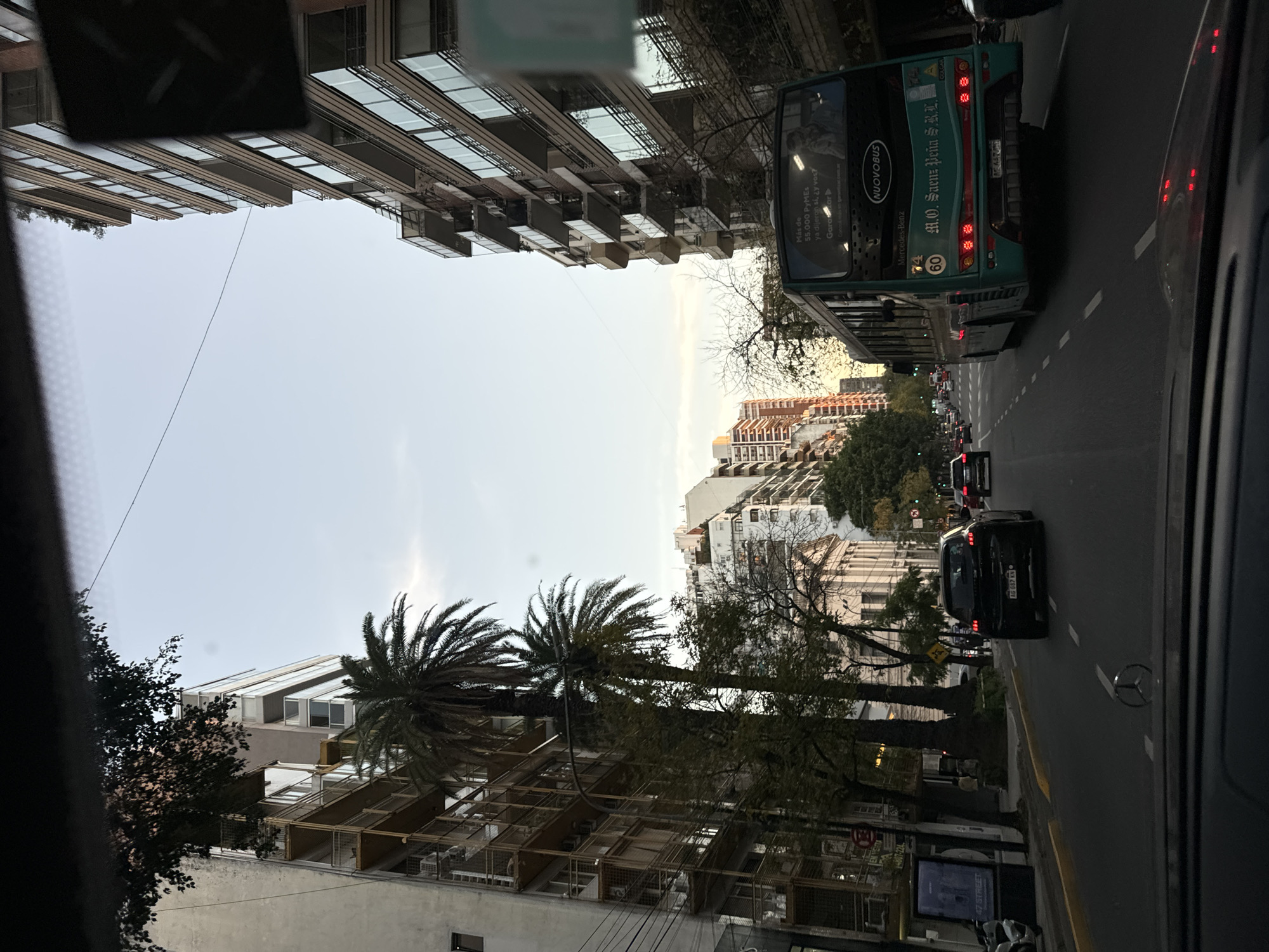 The image shows a street scene in Buenos Aires, Argentina, taken from inside a vehicle. On the right side of the frame, a dark-colored car, a black Mercedes, is driving in the street, a large green and blue bus alongside it. The bus has the words "M.O. Saenz Peña S.K.L." and "Mercedes-Benz" on its rear, as well as "NUOVOBUS". A license plate beginning with "AG" is visible on the car.
The street is lined with trees and buildings on both sides, some of which are high-rise apartments. Palm trees are visible among other trees, adding to the cityscape. A sign is seen advertising "47 Street." The light is muted, suggesting it could be early morning or late afternoon. Other cars are present in the distance, indicating traffic. The sky is overcast.