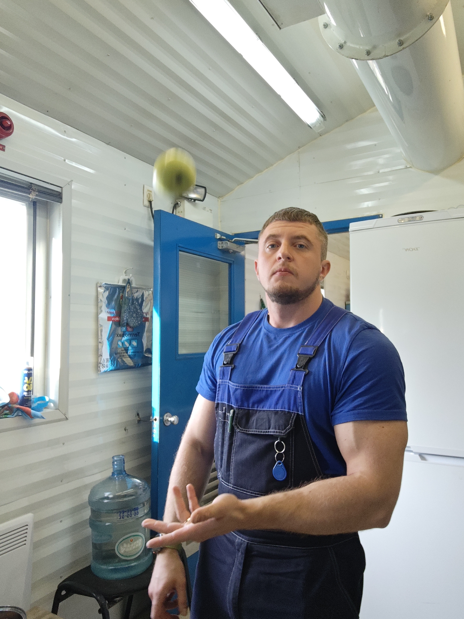 A man in work overalls is captured in what appears to be a functional indoor space, possibly a break room or utility area within a portable cabin or industrial setting. The man, with short brown hair and a beard, looks directly at the camera with a neutral to slightly serious expression. He wears a blue t-shirt and blue and dark grey bib overalls, featuring pens in a chest pocket and a blue key fob on a ring. His left hand is open and slightly raised, while a blurred yellow-green object, possibly a ball or fruit, is visible in mid-air above his right shoulder, suggesting an action shot of throwing or catching.
The setting is characterized by white corrugated metal walls and ceiling, illuminated by fluorescent light fixtures. A partially open blue door is behind the man, and a window with a partially lowered roller blind is on the left. Below the window, a white radiator and a blue spray can are visible. A large, clear plastic 5-gallon water bottle with water sits on a dark stool in the foreground, displaying the numbers "33-31-50" and "20-80-03". A silver insulated bag, with the Cyrillic word "Термобок" (Thermobox) partially visible, hangs on the wall. A red fire alarm is mounted high on the wall above the window. A white refrigerator or freezer, possibly of the brand "YORK", is positioned on the right side of the frame. The bright interior lighting suggests it is daytime.