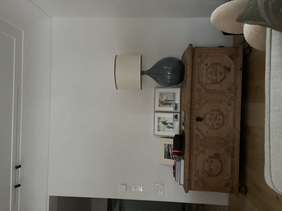 The image shows a wooden chest with intricately carved details, situated in front of a white wall with a single lamp. The chest is positioned in a room with a white ceiling featuring two black track lights. The lamp has a cream-colored cylindrical shade with a brown trim and a dark blue glass base. On top of the chest are three framed pictures depicting people in wintery scenes. There are two white light switches on the wall. There is a hat on top of the chest and several books stacked behind it. The room also includes the partial view of a cream-colored sofa with a white cushion and a green pillow. The floor is wooden and the wall is painted white. The room is located in Celerina/Schlarigna, Switzerland. The coordinates provided are 46.514 latitude and 9.859 longitude.