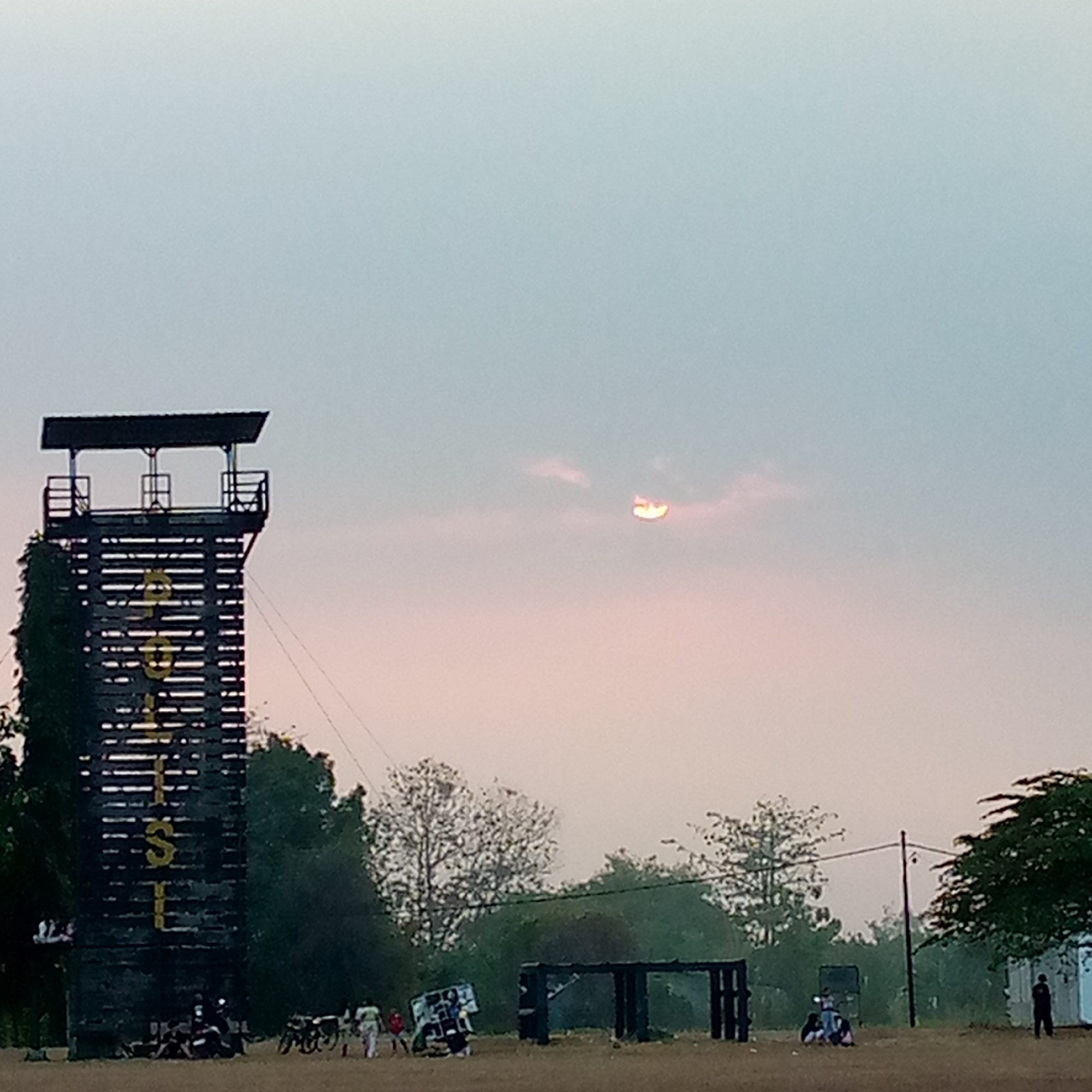 The image depicts an outdoor scene at dusk, likely in Pacing, Indonesia, given the context. A prominent feature is a tall, dark wooden tower structure on the left, with horizontal slats and vertical yellow lettering that appears to spell "POLISI". The tower has a roofed top with railings. In the sky, a partial sun is visible, partially obscured by clouds, creating a soft, diffused light.
The foreground consists of a dusty or dirt field. To the left of the tower, several people are gathered, some near what appears to be motorcycles. Further to the right, there are more individuals walking and standing, along with several bicycles. In the middle ground, there's a dark, rectangular structure resembling an open frame or gate. Trees are scattered throughout the scene, with denser foliage behind the tower and on the right side. A power pole is visible on the right, with wires extending across the image. The overall atmosphere is quiet and still, characteristic of early morning or late evening.