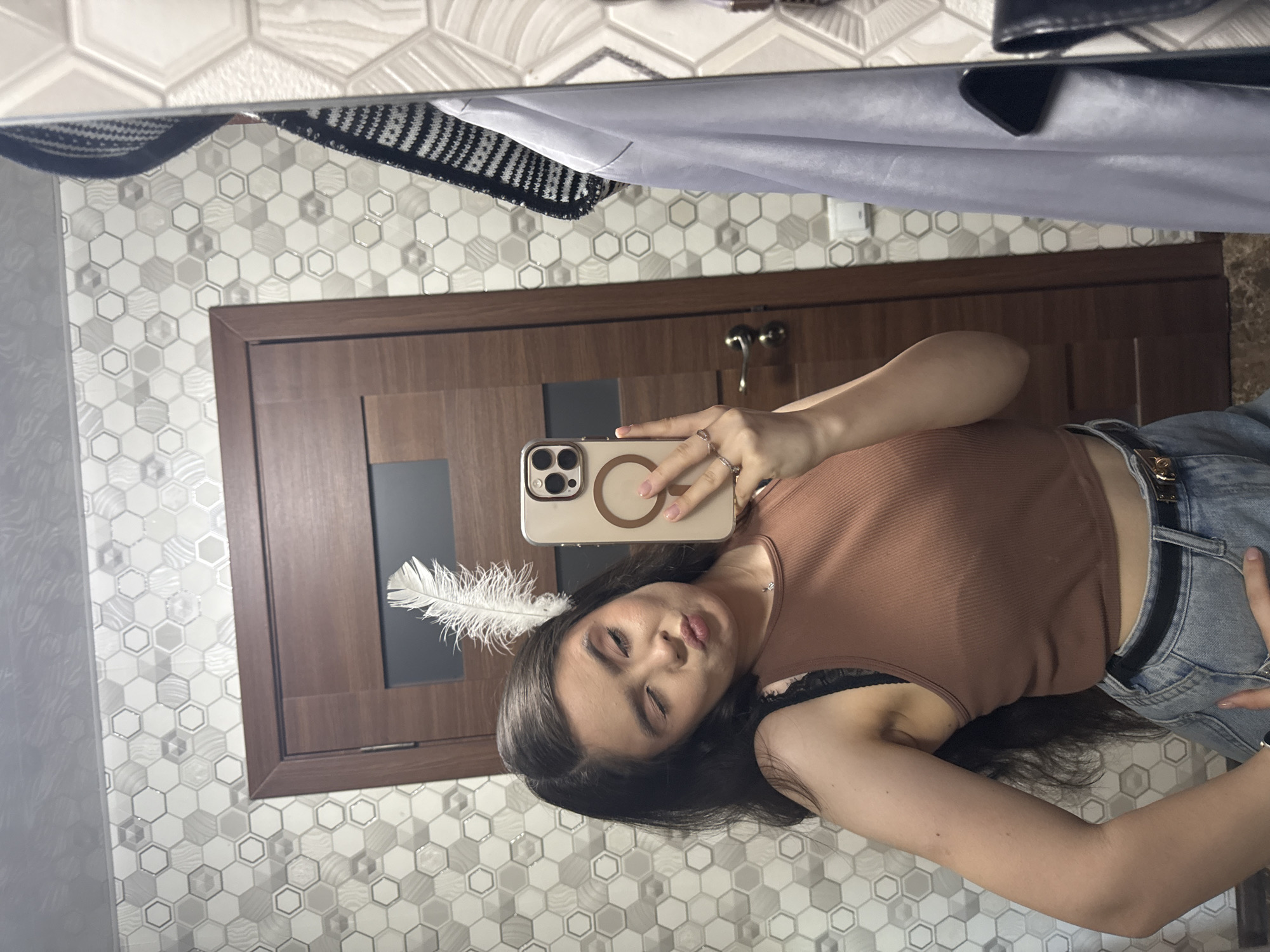 A young woman is taking a mirror selfie in what appears to be a bathroom or dressing area. She has long, dark hair and is wearing a brown crop top, a black bra strap visible underneath, and denim shorts with a black belt. She is playfully winking and puckering her lips. A white feather is tucked behind her ear. She is holding a smartphone with a beige case in her right hand to capture the reflection. The background shows a tiled wall with a hexagonal pattern in shades of white and gray, a wooden door, and a closet with clothing hanging on the right side. The overall scene is brightly lit and suggests a personal, casual moment. There are no indications of the location being in Shulhynka, Ukraine.