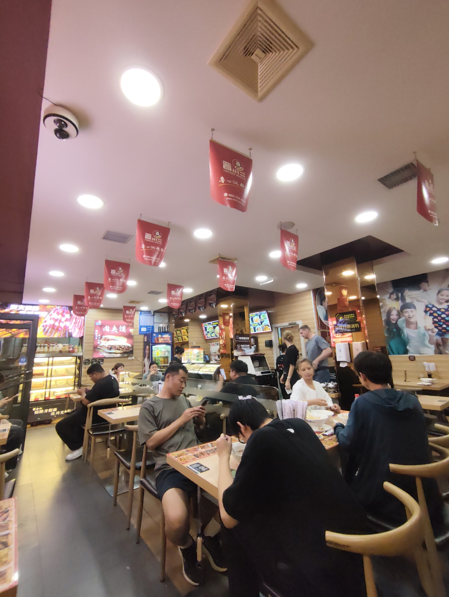 The image depicts the interior of a restaurant, possibly a noodle shop, located in Aihui, China. The scene is busy with customers seated at tables and staff attending to them. The decor features red banners hanging from the ceiling with Chinese characters. Various food items and displays are visible, including what appears to be a refrigerated display case filled with food. In the foreground, several individuals are seated at tables, some engaged with their phones, while others are eating. The lighting is bright, primarily from overhead circular fixtures. The overall atmosphere suggests a casual dining environment.