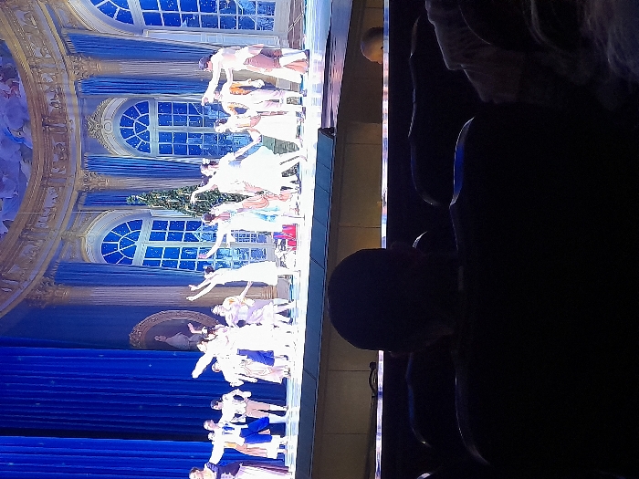 A ballet performance unfolds on stage with dancers in white dresses and blue accents. Behind them, a painted backdrop features arched windows with starry designs and blue curtains. Audience members are visible in the foreground, silhouetted against the performance. The scene is illuminated by stage lights, creating a vibrant and captivating atmosphere.