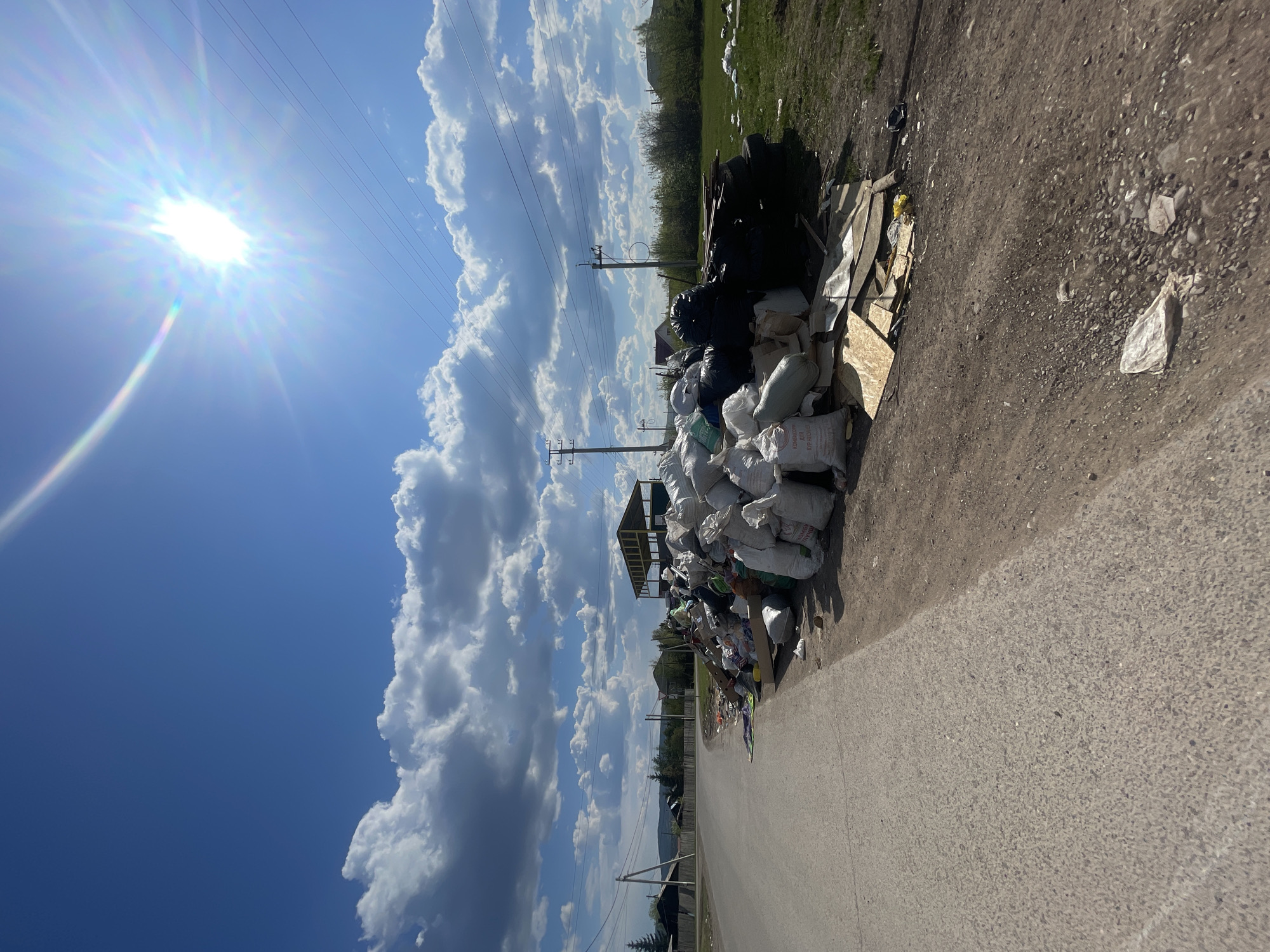 The image shows a large pile of garbage bags and debris along a road in Минино, Russia. The garbage includes white sacks, black plastic bags, tires, plywood, and assorted trash. A road runs along the right side of the pile. In the background, power lines stretch across a blue sky with large white clouds. The sun is brightly visible in the upper left corner of the image.