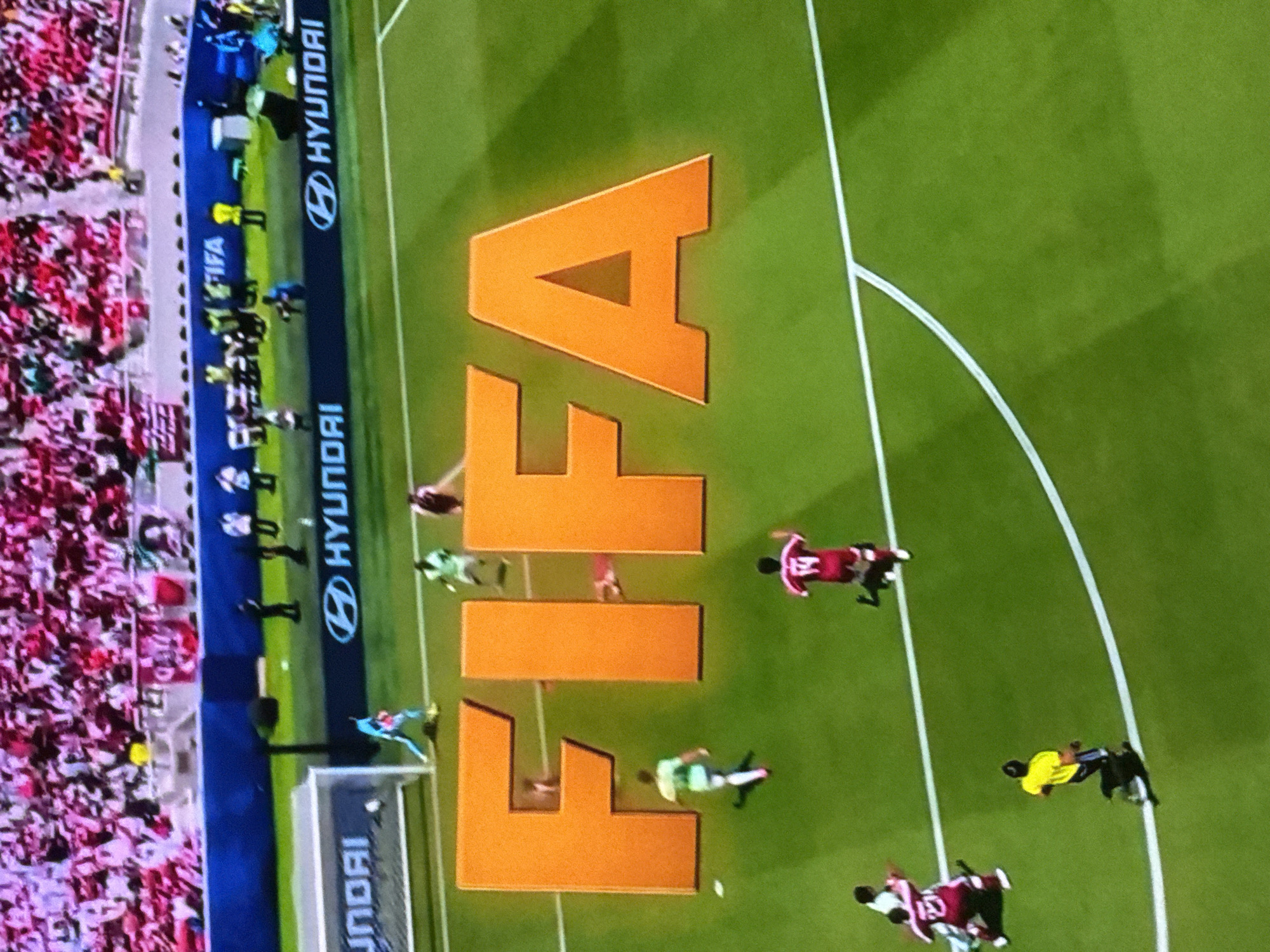 An aerial view of a soccer match, with a large orange "FIFA" logo prominently displayed on the green grass field. Several soccer players in various colored uniforms are scattered across the field, some in motion, suggesting an active game. In the background, a stadium filled with spectators wearing pink and white attire is visible, along with advertising banners along the perimeter of the field, including "HYUNDAI." The scene appears to be an outdoor sporting event.