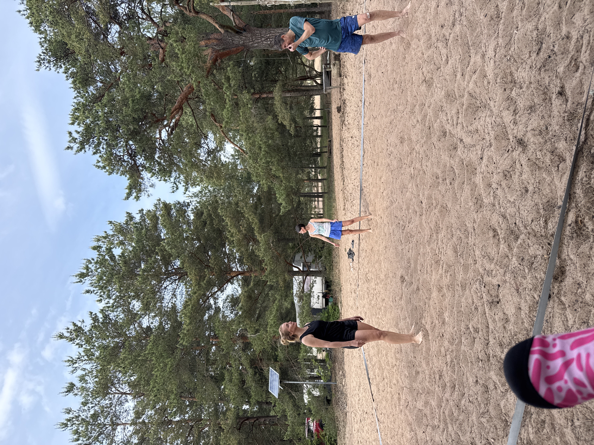 This image captures an outdoor scene at Peski, Russia, featuring three individuals on a sandy beach volleyball court surrounded by a pine forest under a partly cloudy sky. In the foreground, at the bottom left, a foot wearing a pink sock with a black toe is visible, indicating the viewer's perspective. A dark grey court boundary tape stretches across the mid-ground.
Three adults are positioned on the sand. On the left, a blonde woman stands barefoot, wearing a black tank top and black shorts, looking towards the center-right. In the center-right, a dark-haired woman stands barefoot, wearing a light-colored patterned tank top and blue shorts, facing towards the left. On the far right, a man with short dark hair stands barefoot, wearing a dark green t-shirt and patterned blue shorts. A light-colored bandage is visible on his left knee. He has his right hand raised towards his face and his left hand on his hip, appearing to be in mid-reaction or communication. A wooden net post for the volleyball court is visible behind him.
The background consists of a dense pine forest. Among the trees on the left, a small solar panel mounted on a pole is visible, along with a partial view of a red vehicle and a white recreational vehicle or trailer further back. The ground is consistently sandy with visible undulations and footprints. The sky overhead is light blue with scattered white clouds, indicating an overcast or partly cloudy daytime setting.
