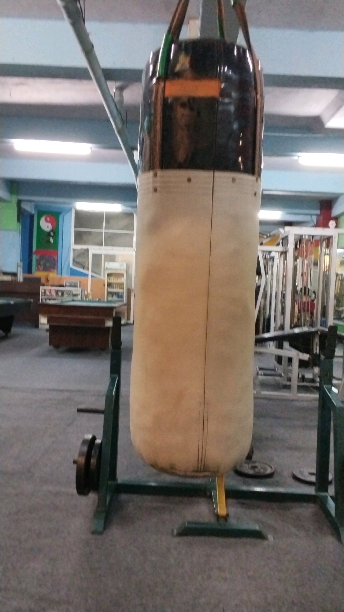 A tall, beige punching bag hangs from the ceiling in the foreground of the image. It is suspended by straps from an overhead metal support. The top portion of the punching bag is dark, possibly leather or a similar material, with reflective panels. The bag is supported at the bottom by a green metal stand. To the left of the punching bag, a weight plate is attached to the stand. In the background, the scene appears to be an indoor sports facility or gymnasium. There are various pieces of gym equipment, including weight machines, visible. In the far background, there are windows and doors, and on the left wall, a colorful depiction of the Yin and Yang symbol is visible. The floor is carpeted in a dark gray color. The lighting suggests fluorescent overhead lights. The scene is static and shows no people or any ongoing activity. The location is identified as Sentanan, Indonesia.