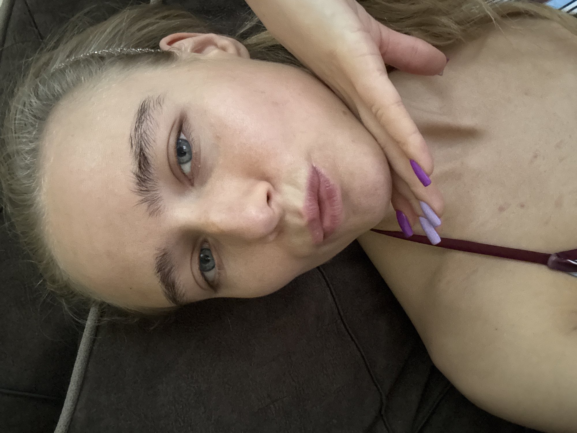 A fair-skinned young woman with blonde hair is lying on a dark brown couch. She has blue eyes and prominent eyebrows. Her lips are pursed. Her right hand is near her face, with purple and light purple nail polish on her nails. She has on a thin, silver-colored headband. A maroon strap is visible near her shoulder. The location and city are unknown.