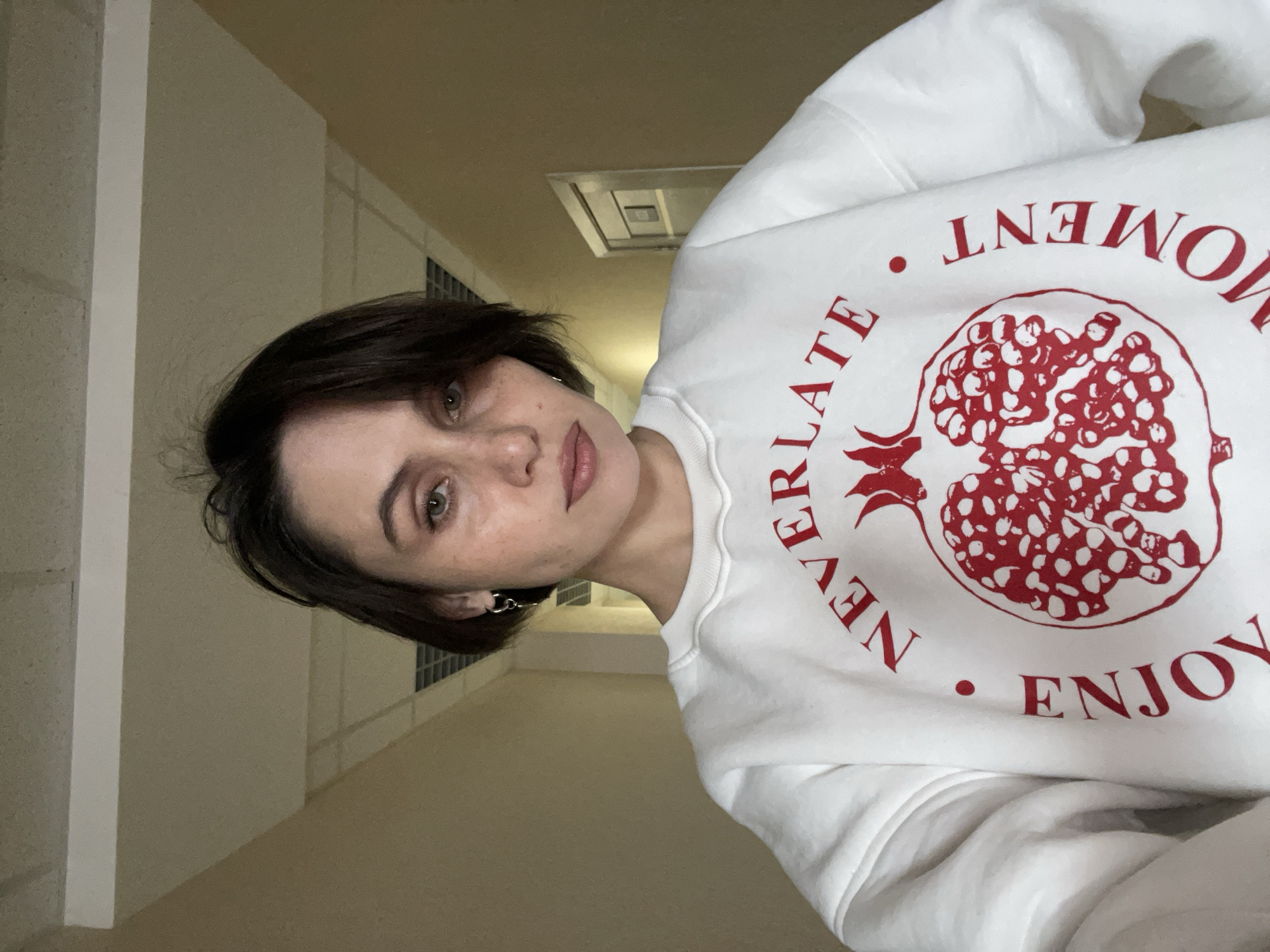 A young woman is captured in a close-up portrait, looking directly at the viewer with a neutral expression. She has short, dark brown hair and light-colored eyes. She is wearing a white sweatshirt featuring a large red illustration of a pomegranate. Red text curves around the design; "NEVERLATE" is visible along the left side, and "ENJOY" is along the lower right. Another word, appearing partially upside down as "TN∃WON" in the image, which would spell "MOMENT" when viewed upright, is visible at the top right.
The background suggests an indoor setting, likely a hallway or corridor, characterized by plain, light-colored walls and a matching ceiling. A rectangular vent or light fixture is visible on the ceiling. The lighting appears artificial, indicating an indoor environment, possibly at any time of day. The scene is devoid of specific decorative details, emphasizing a functional space in Izhevsk, Russia.