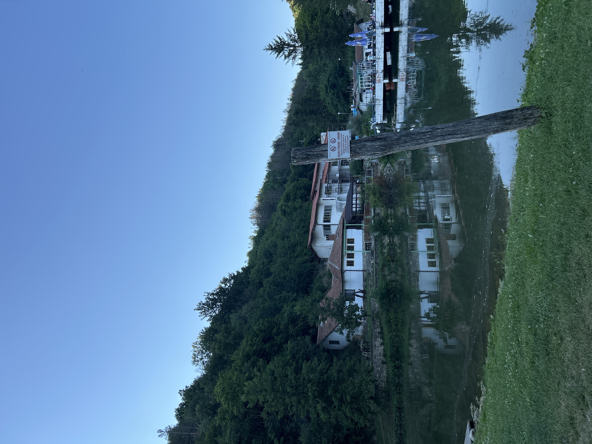 The image depicts a waterside scene with buildings nestled into a forested hillside, reflected in the calm water. On the left, a dense green forest covers a steep incline. Several white buildings with red tile roofs are visible through the trees, appearing to be built into the hillside. One building has a large open area with white tables and chairs. Further up the hillside and to the right, another structure with a flat roof and white railings extends over the water, with umbrellas set up and people visible on its deck. A weathered wooden post stands vertically in the foreground, jutting out of the water. A sign is attached to this post. The water reflects the buildings and trees, creating a mirrored image. The right side of the image shows a grassy bank. The sky is clear and blue. The overall scene suggests a tranquil location, possibly a resort or restaurant area by a lake or river.
The sign on the wooden post contains text in what appears to be a Cyrillic alphabet. The visible text includes "ЗАБРАНЕНО" (Forbidden) at the top, followed by a warning symbol with a prohibition sign. Below this, there are further lines of text which are difficult to read clearly but appear to detail specific prohibitions, possibly related to swimming or entering the water.
The time of day appears to be late afternoon or early evening, given the angle of the light and the clear blue sky. The weather seems pleasant. The location is not explicitly identified but evokes a natural, possibly recreational, setting.