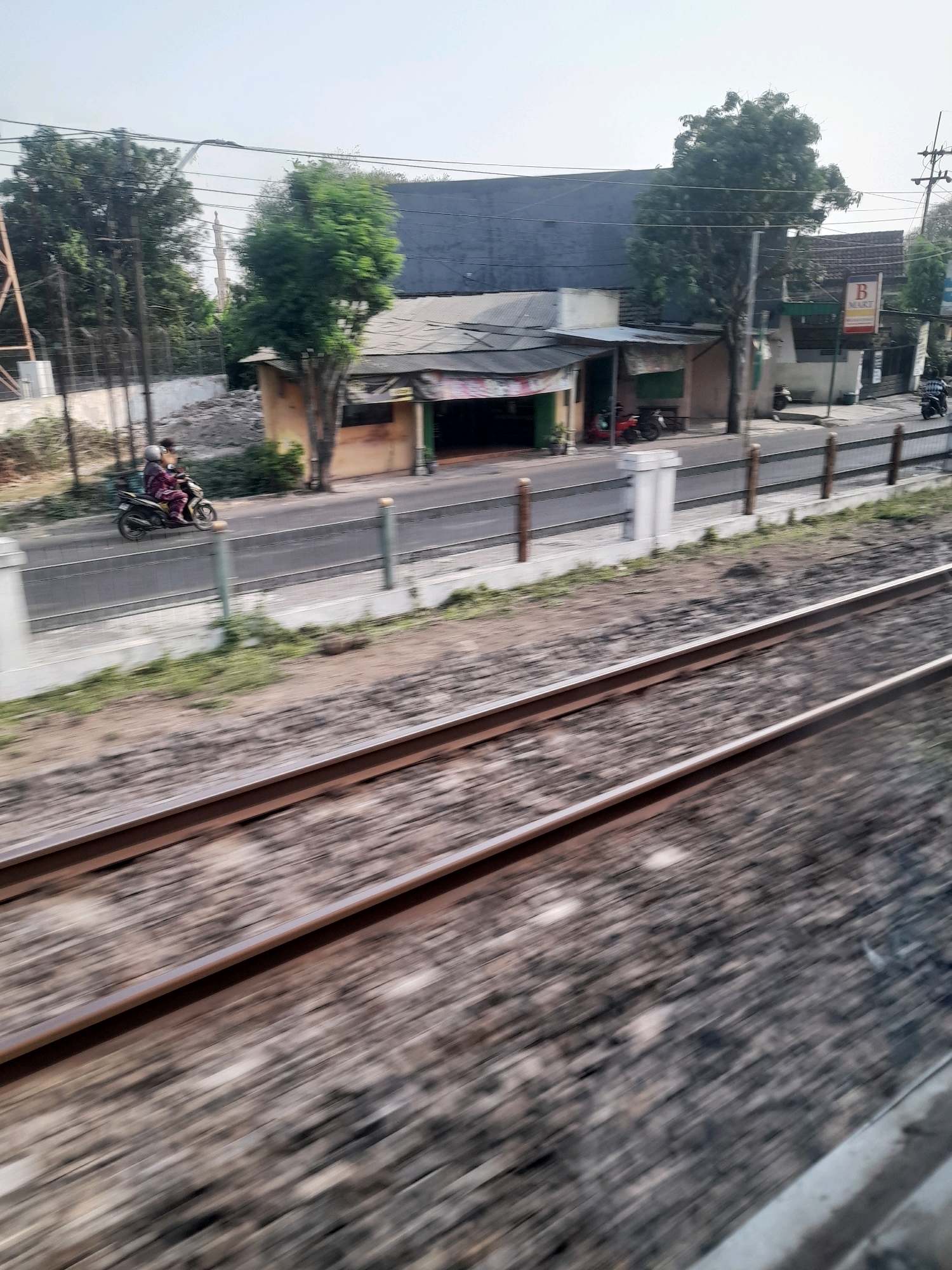 An eye-level, medium shot captures a street scene in Peterongan, Indonesia, as viewed from a moving train. In the foreground, blurred train tracks dominate the lower half of the frame. The middle ground features a paved street with a white concrete barrier and wooden posts separating it from the tracks. A person rides a motorcycle wearing a maroon outfit and a helmet, moving along the street. Along the street, a small building with a corrugated metal roof and an open front appears to be a shop. Next to it, another building has a sign with the letter "B" and the word "MART" visible. Beyond the buildings, trees and utility poles are present. The background consists of a hazy sky.