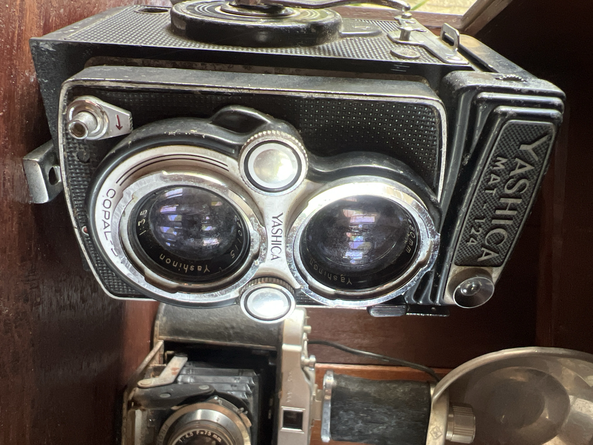 This is a close-up, eye-level, medium shot of a vintage Yashica MAT 124 twin-lens reflex camera. The camera is black and silver, with prominent lenses and controls. The branding "YASHICA" is visible on the camera body and lenses. A flash unit is attached to the top right of the camera, and another older camera is visible in the background. The cameras are positioned on a wooden surface, likely a shelf or table, with a dark, out-of-focus background that suggests an indoor or shaded environment. The scene is well-lit, highlighting the details and textures of the vintage equipment. The location is identified as Ubud District, Indonesia.