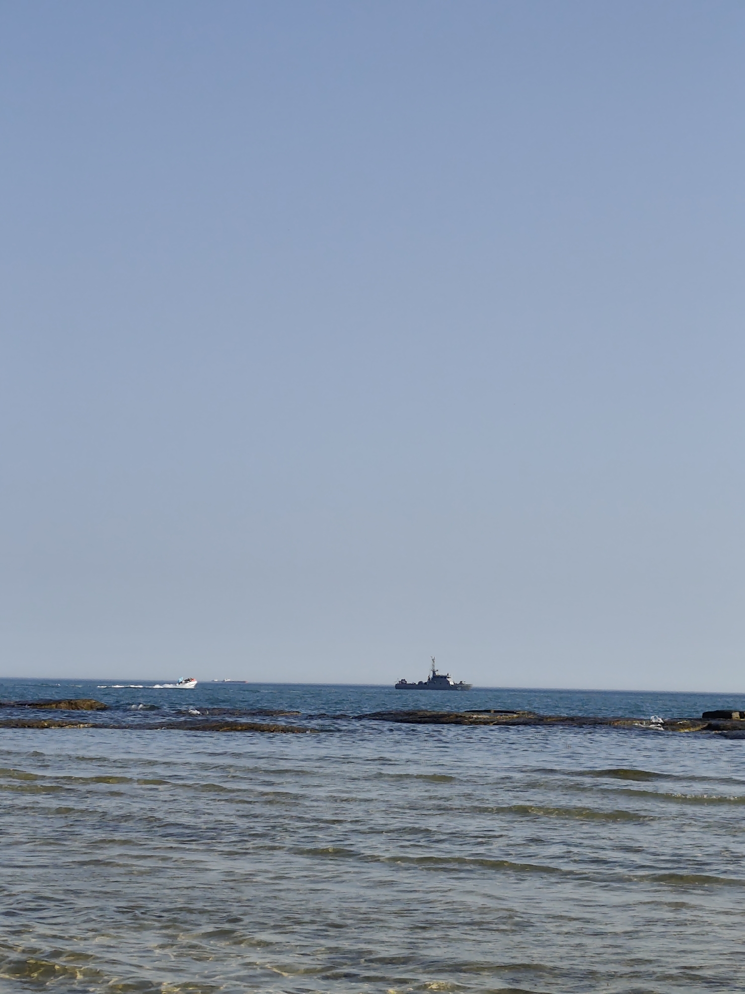 The image captures a tranquil scene of the Caspian Sea near Aktau, Kazakhstan, under a clear, pale blue sky, suggesting a warm, sunny day with calm weather. The foreground is dominated by the gently rippling surface of the water, where sunlight glints off the waves, revealing the sandy or rocky seabed beneath in clearer areas. A line of weathered rocks, partially submerged, stretches across the middle ground, breaking the expanse of the sea.
Further out in the distance, two vessels are visible. To the left, a small white motorboat with at least two people aboard is creating a white wake as it moves across the water. To the right, a larger, dark-colored ship, possibly a patrol or naval vessel, stands out against the horizon. Its silhouette is distinct, with a tall structure at the rear. The overall mood of the image is one of peaceful serenity, with the vastness of the sea and sky creating a sense of calm and openness. The time of day appears to be late morning or early afternoon, given the bright sunlight. There is no visible text in the image.